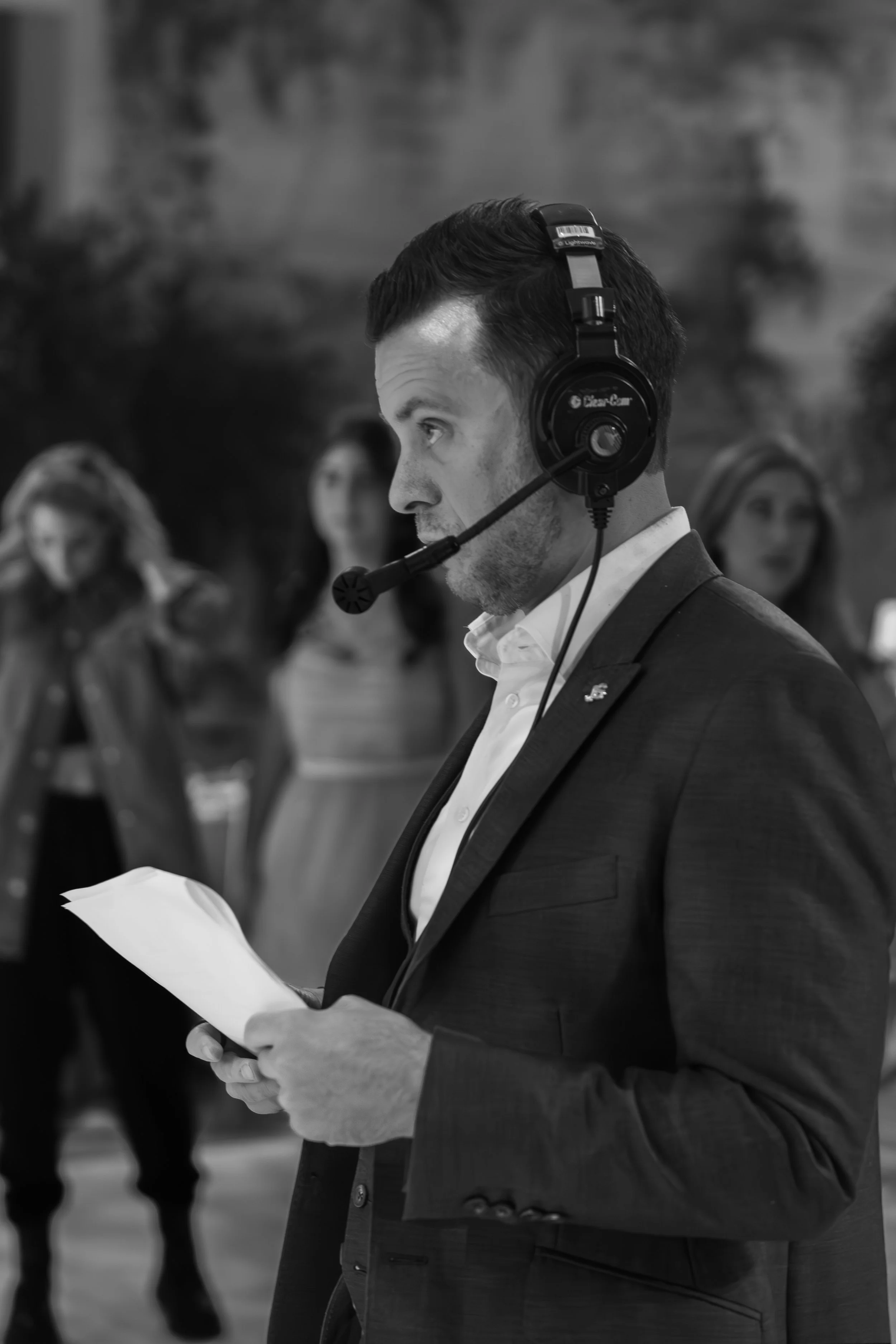 Laurie Weitzkorn in a suit wearing a headset and looking at papers, standing indoors with a blurred background of other people.