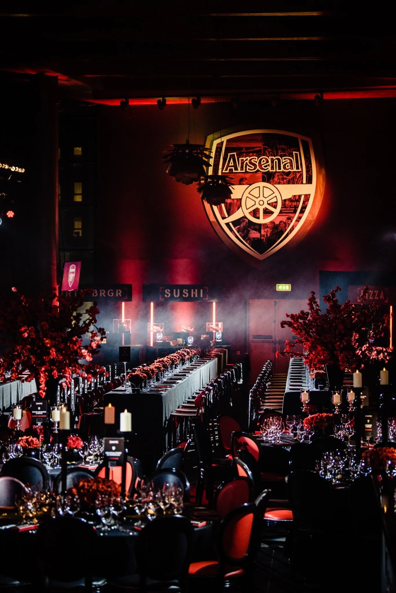 A banquet hall decorated for a formal event with tables adorned with red flowers, candles, wine glasses, and place settings. The hall features a large Arsenal football club logo on the wall and red lighting.