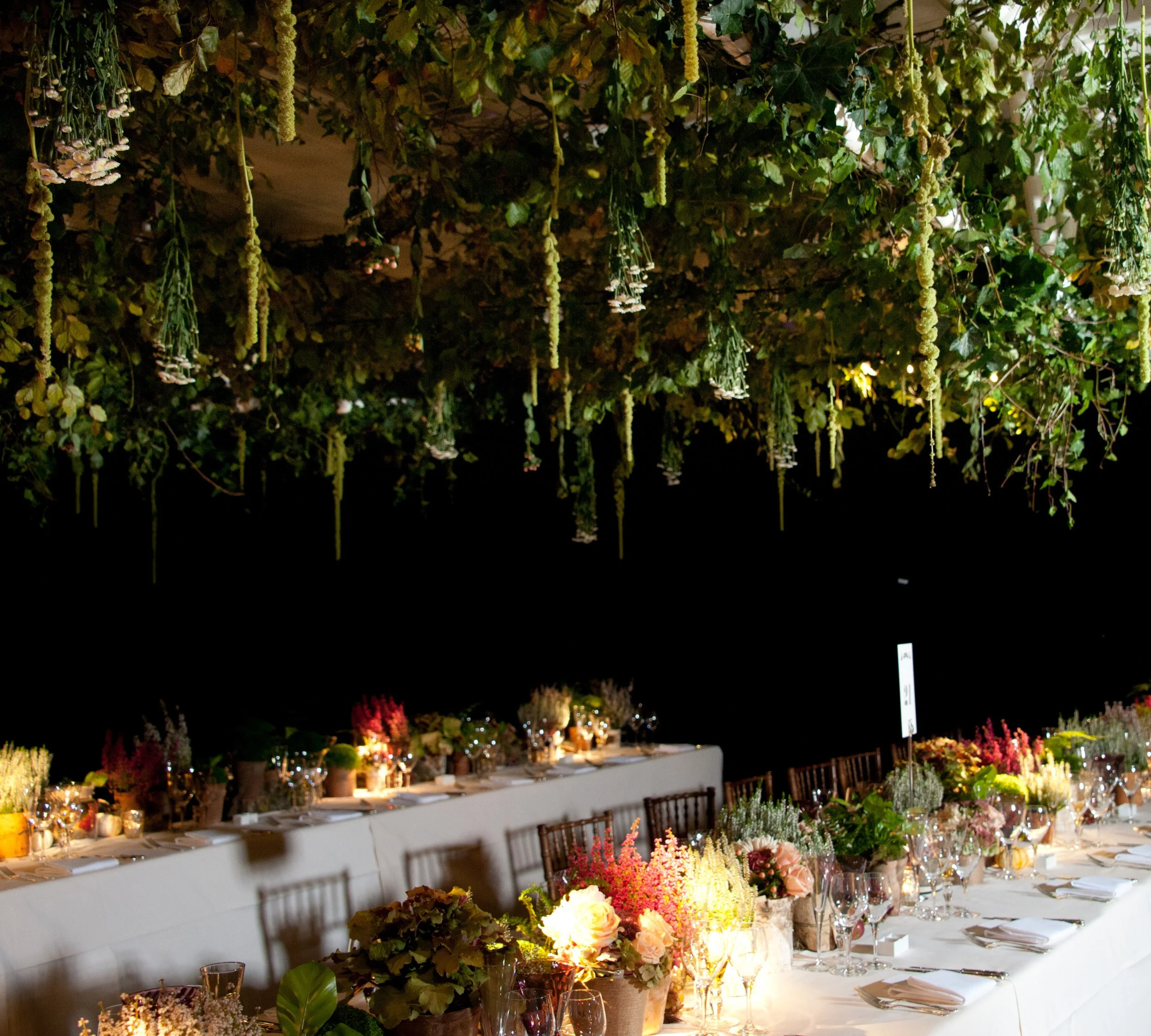 A beautifully decorated indoor dining area set for an event with long tables covered with white tablecloths, floral centerpieces, and candles, with a lush ceiling of hanging flowers and greenery illuminated by soft lighting.