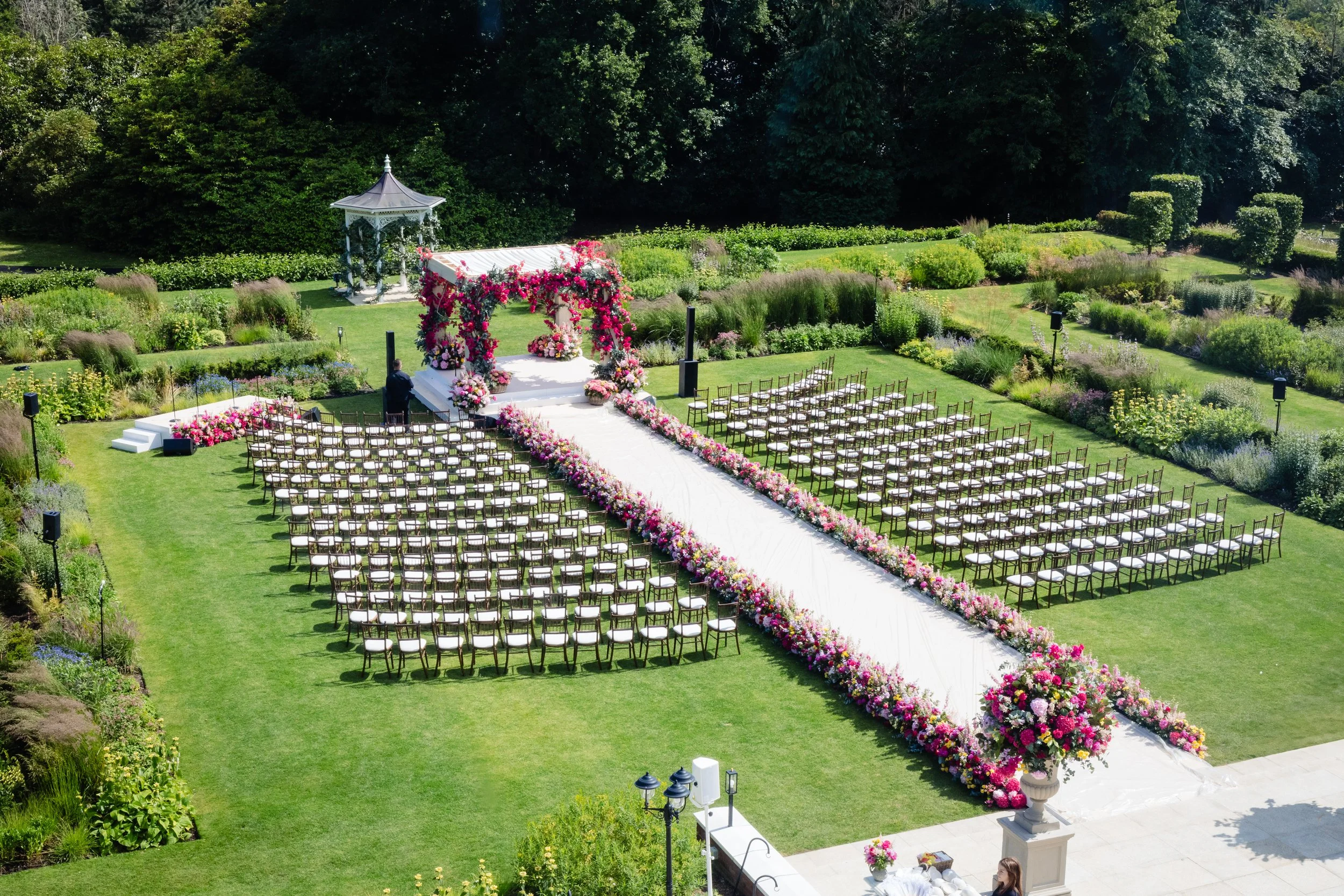 Fairmount House Leatherhead jewish wedding ceremony setup with rows of chairs, a white aisle and floral arch, surrounded by lush greenery and trees and a bright chuppah