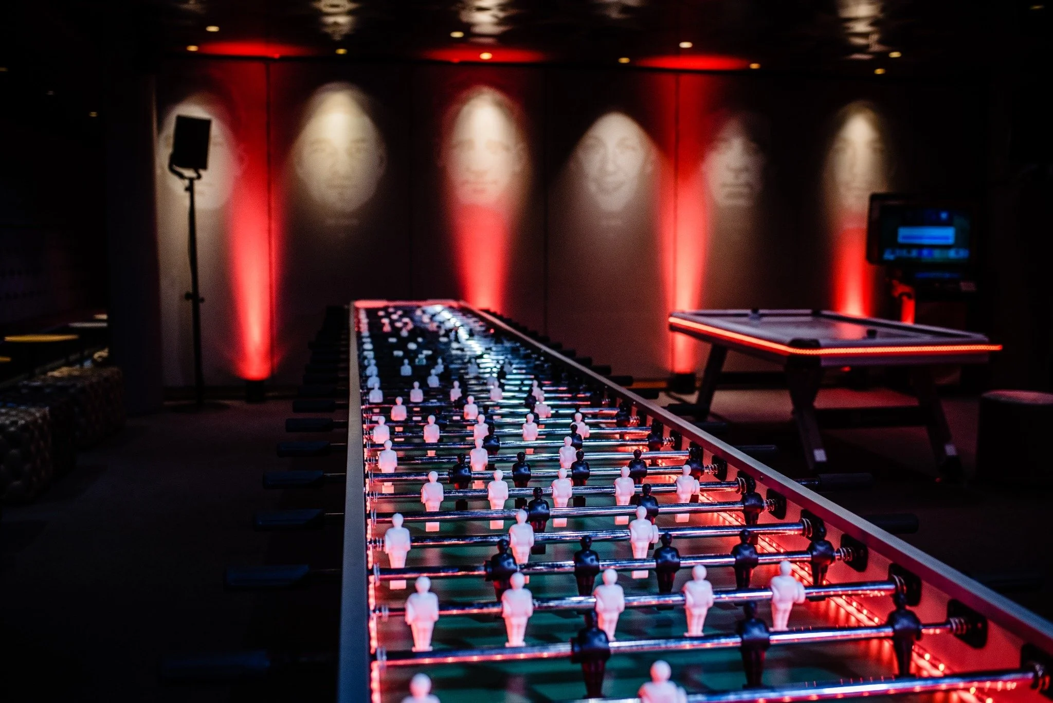 Foosball table with miniature players, illuminated by red and blue lighting in a dark room, with a wall featuring portraits of faces in the background.