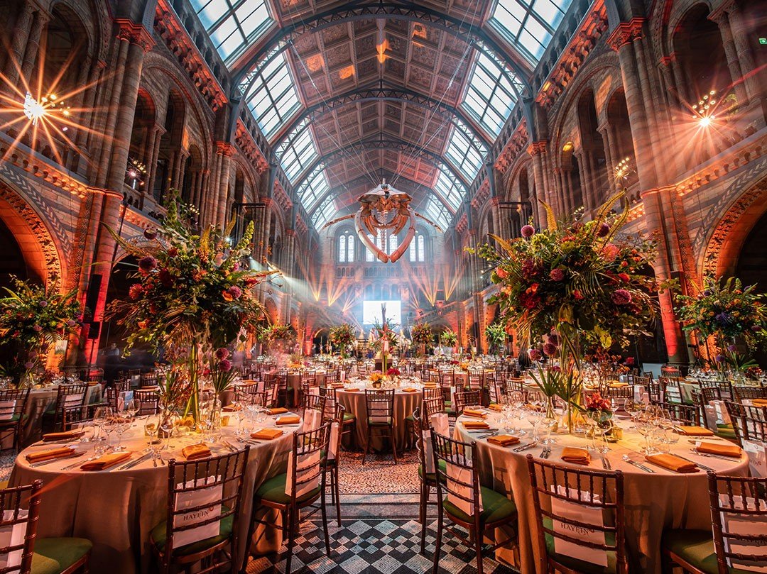 Elegant corporate events night  at The Natural History Museum with high arched glass ceiling, ornate brick walls, and large floral arrangements on round tables set for a formal dinner.