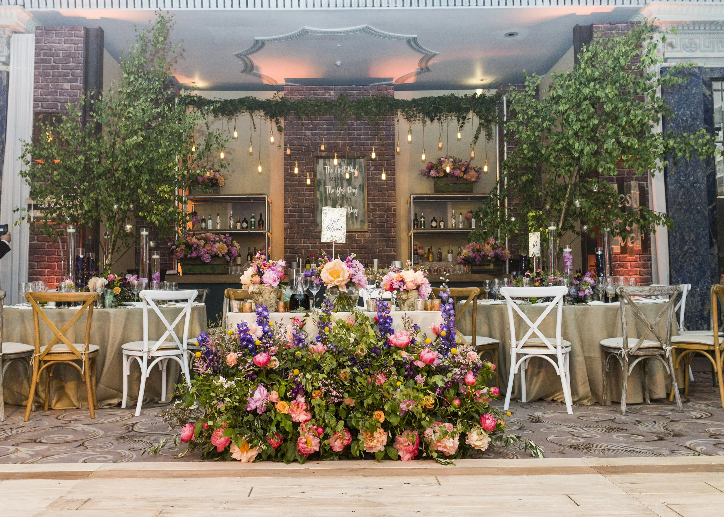 Indoor wedding reception decorated with an abundance of pink, purple, and orange flowers on a long table, set against a brick wall with hanging Edison bulbs and greenery.