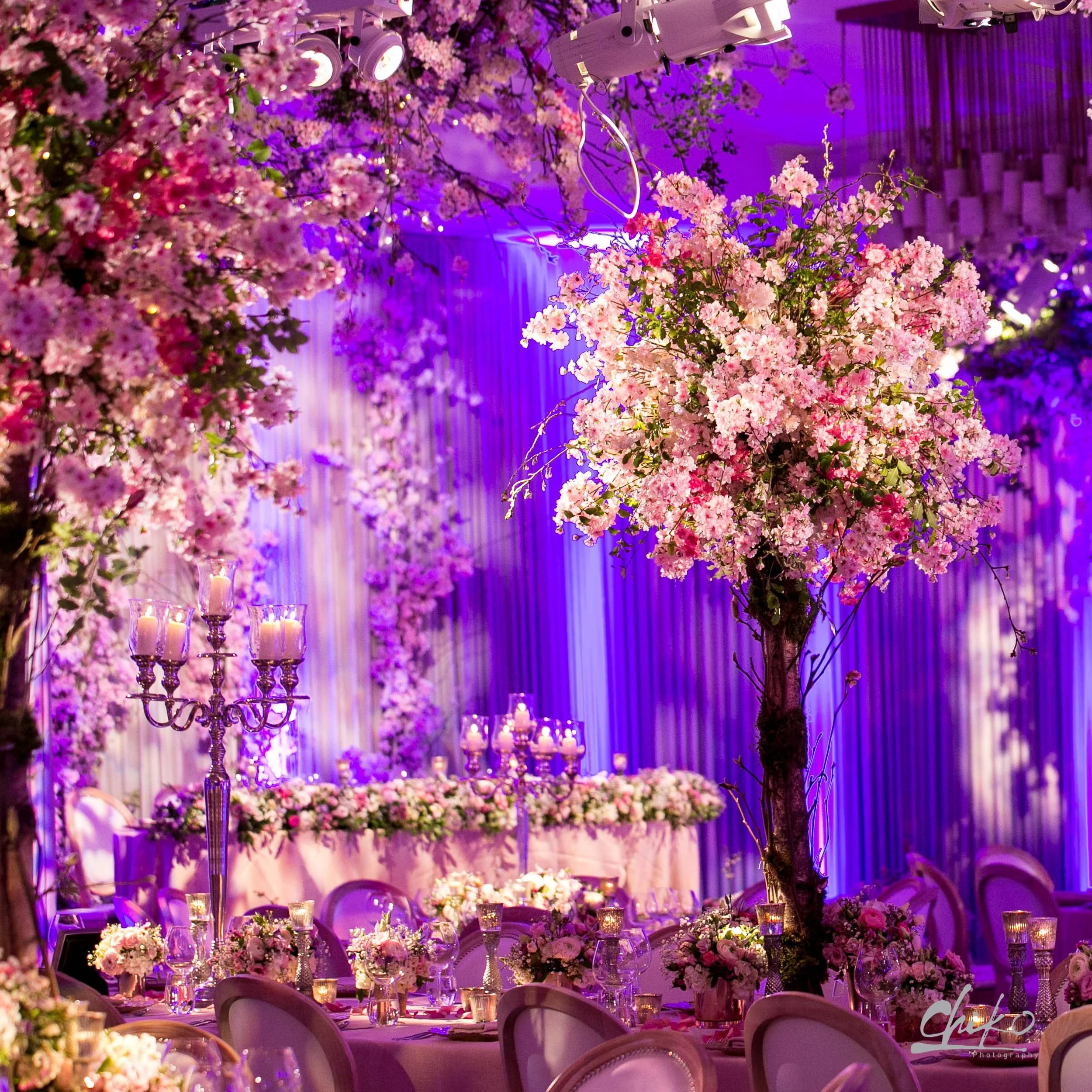 A decorated event space with pink floral arrangements and purple lighting, featuring a long tablescape with candles, glassware, and flower centerpieces, suitable for a wedding or celebration.