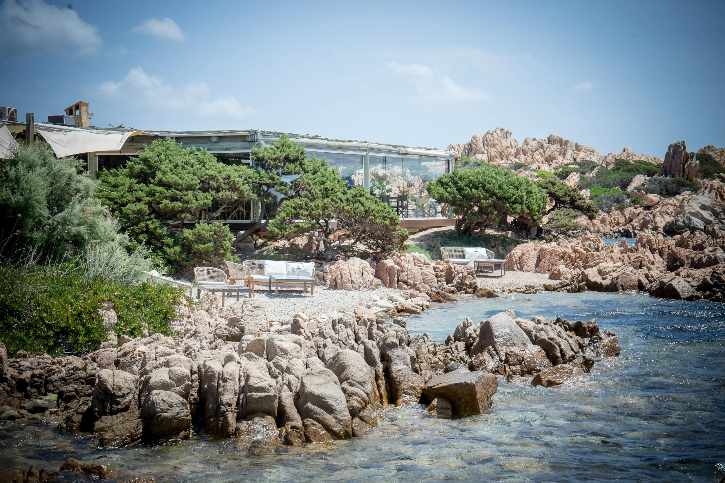 A rocky shoreline with clear water in the foreground, green bushes and trees, a modern glass building with a patio and umbrellas, and a mountainous landscape with blue sky in the background.