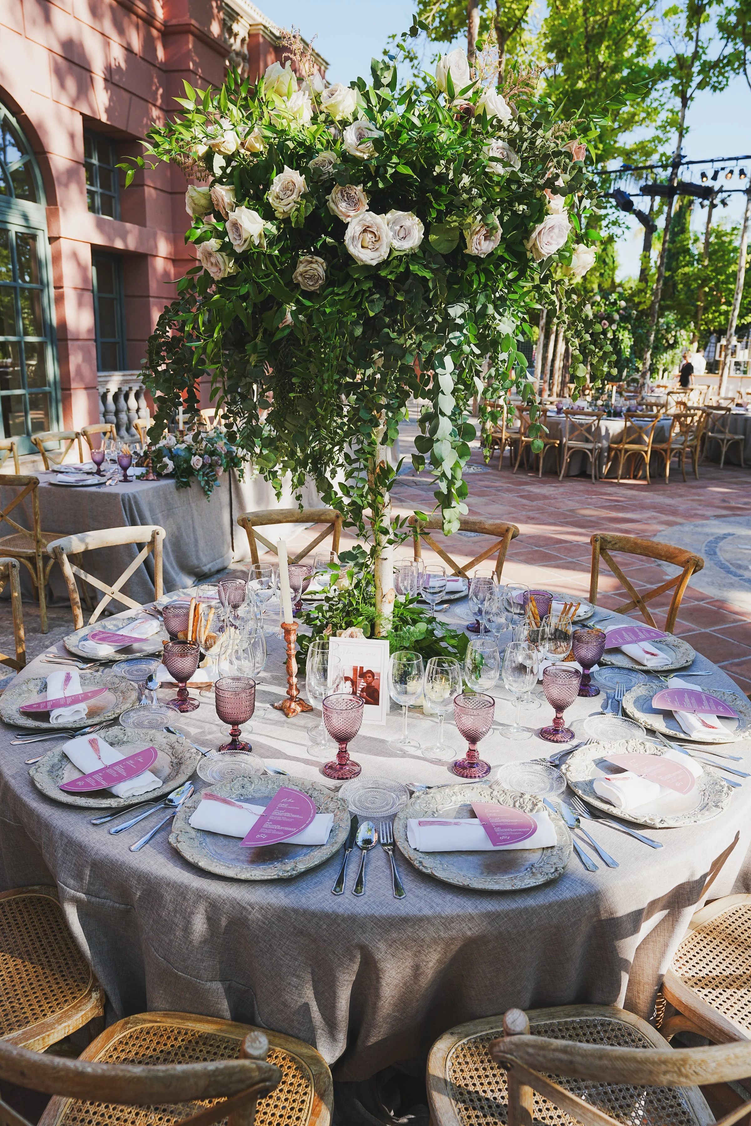 A round dining table decorated with a floral centerpiece, set with plates, napkins, silverware, and purple-tinted glassware, located outdoors with a building, trees, and additional tables in the background at Anantara Villa Padierna Resort 