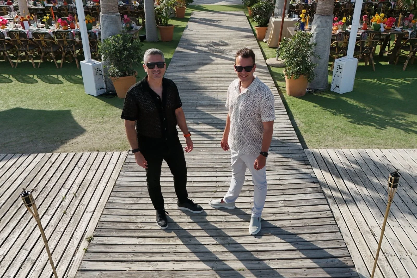 Just Seventy Stas Anastasiou and Laurie Weitzkorn standing on a wooden walkway at an outdoor wedding with tables and chairs decorated with flowers in the background. At Anantara Villa Padierna Resort in Marbella