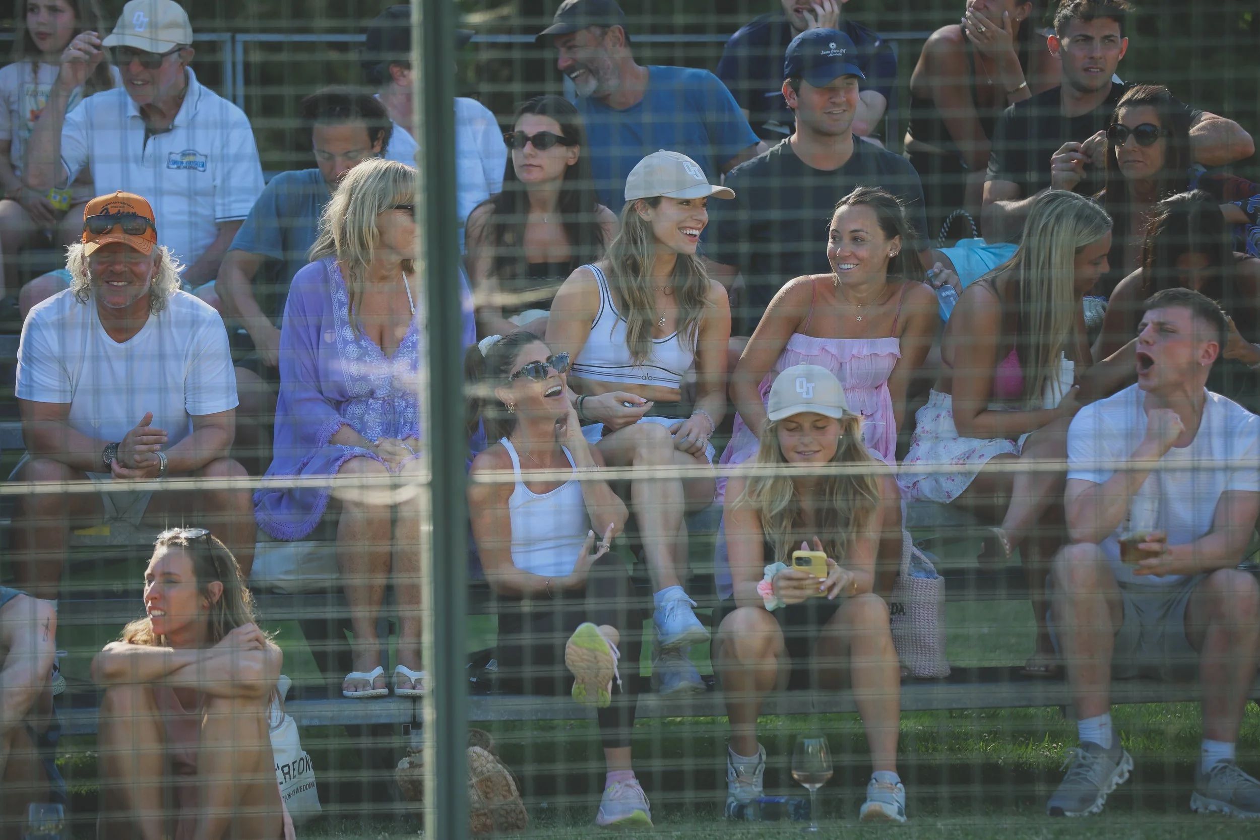 A group of people sitting on bleachers at an outdoor event, seen through glass with reflections, some smiling, talking, and looking at a phone, dressed casually in summer clothing.