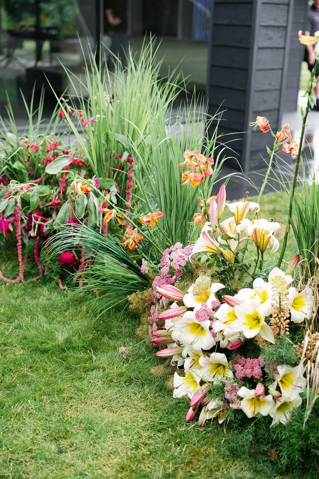 Colorful flower arrangement including lilies, orchids, and pink flowers on lush green grass with a building and glass window in the background.