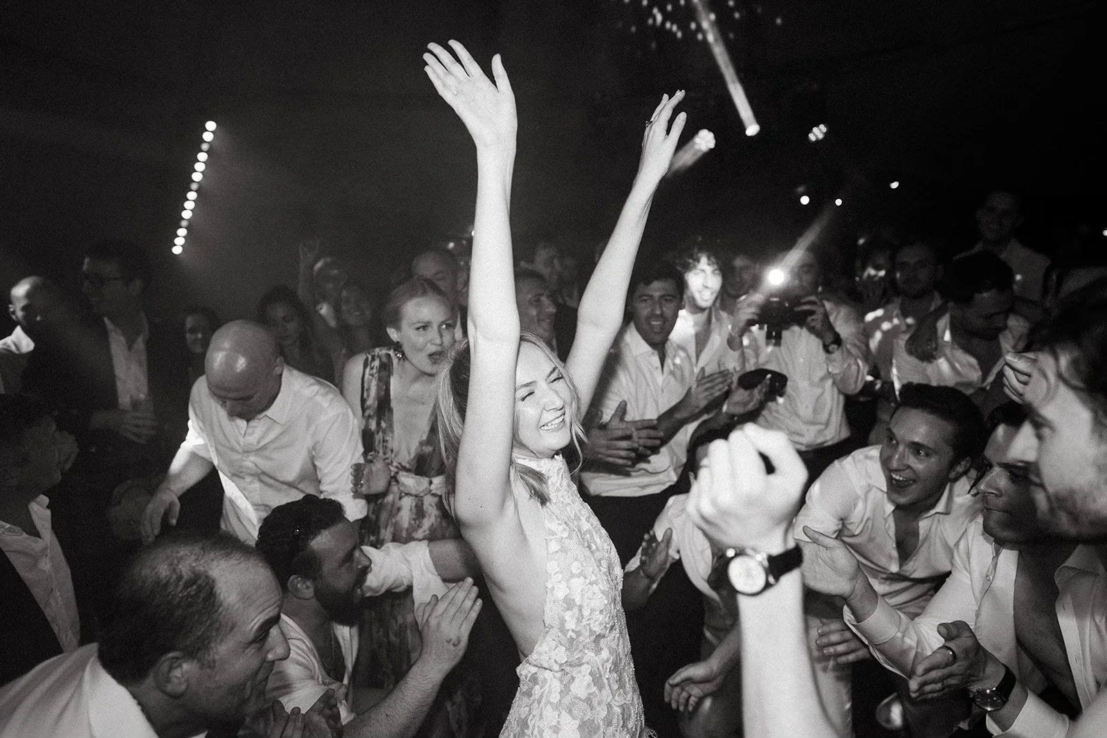 The bride in a sleeveless lace dress is smiling with her arms raised as she dances at a lively afterparty with many people around her. The crowd is celebrating and enjoying the moment, with some clapping and others taking photos.