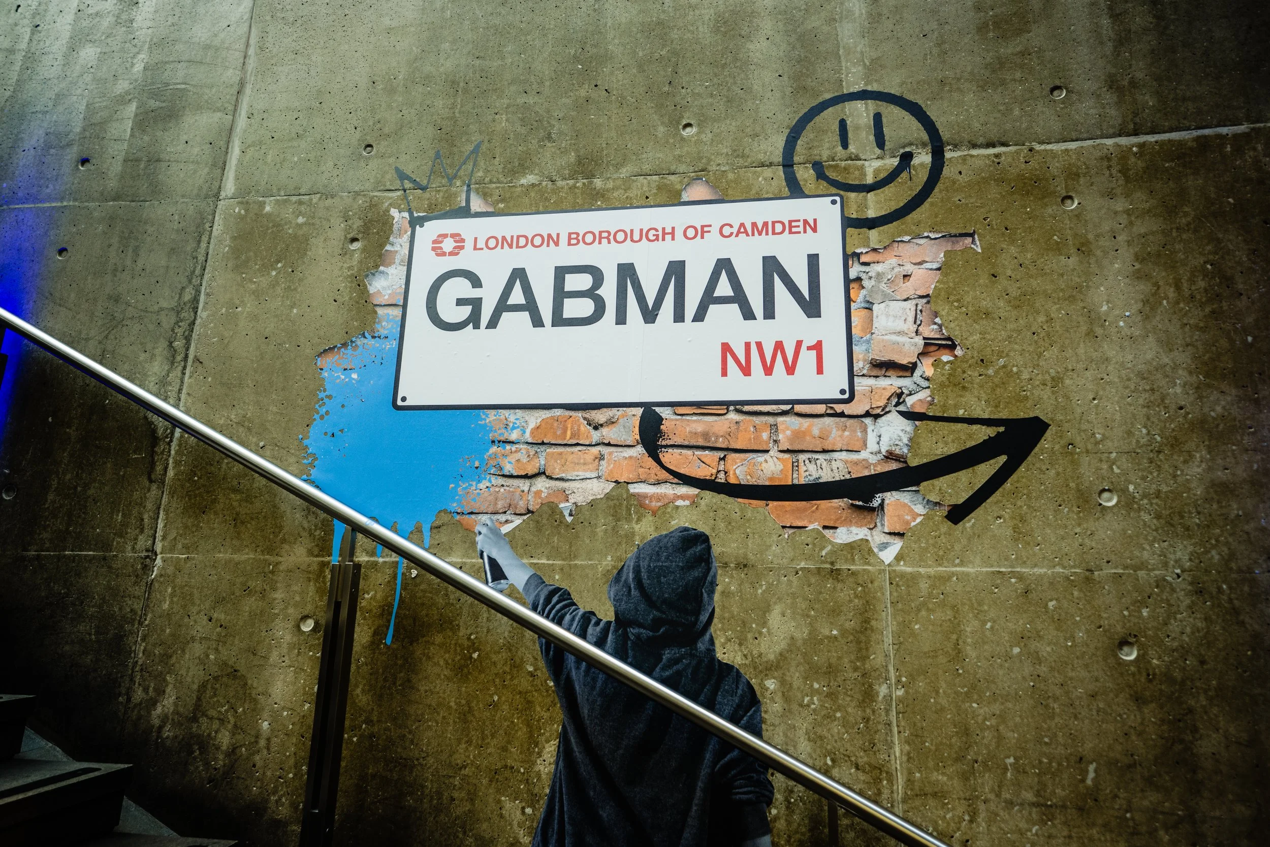 A photo of a street sign for Camden in the London Borough of Camden, with graffiti of a smiling face and a crown, and a distressed mural background on a concrete wall, viewed from above with a person's foot in the bottom center.