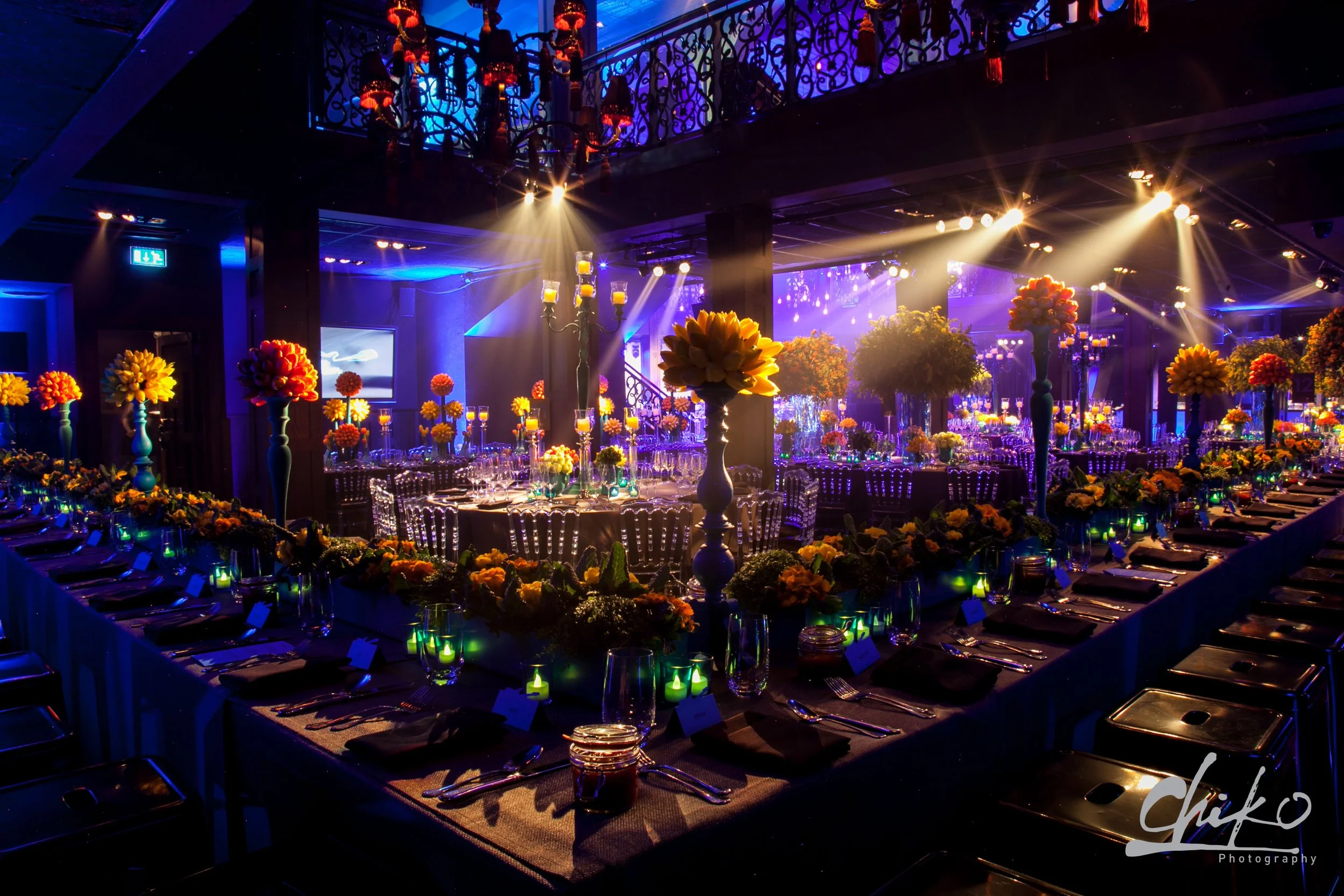 Elegant banquet hall decorated with yellow and orange flowers and candles, illuminated with purple and yellow lighting for a celebration or wedding reception.