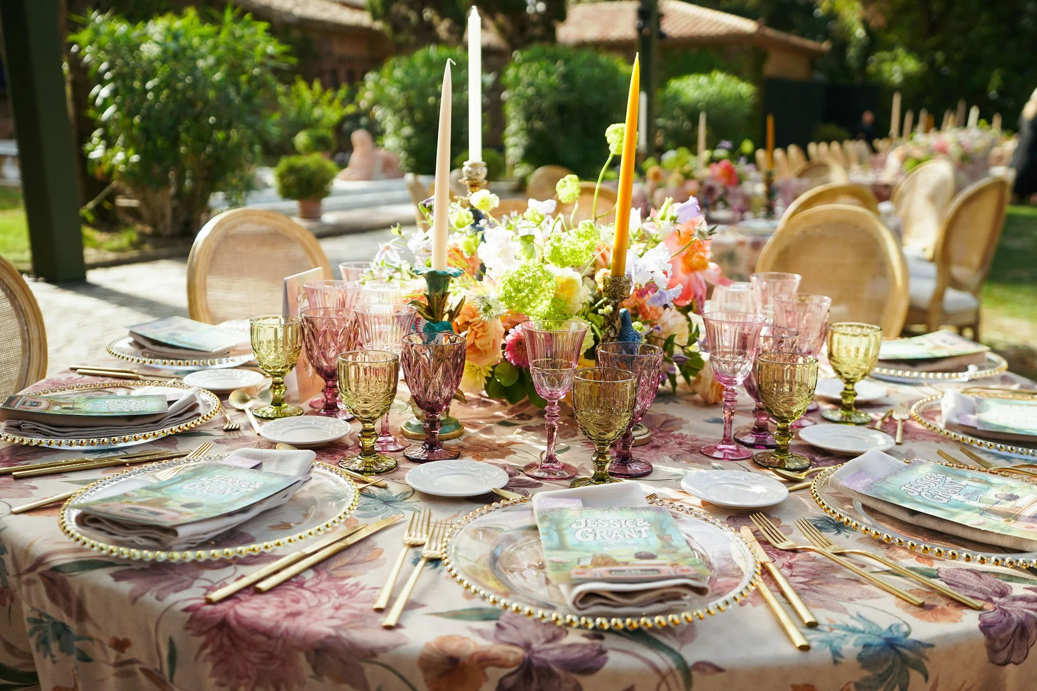 Outdoor event table set with floral centerpiece, candles, pink glassware, gold cutlery, and menus, surrounded by chairs with a lush garden and pavilion in the background.