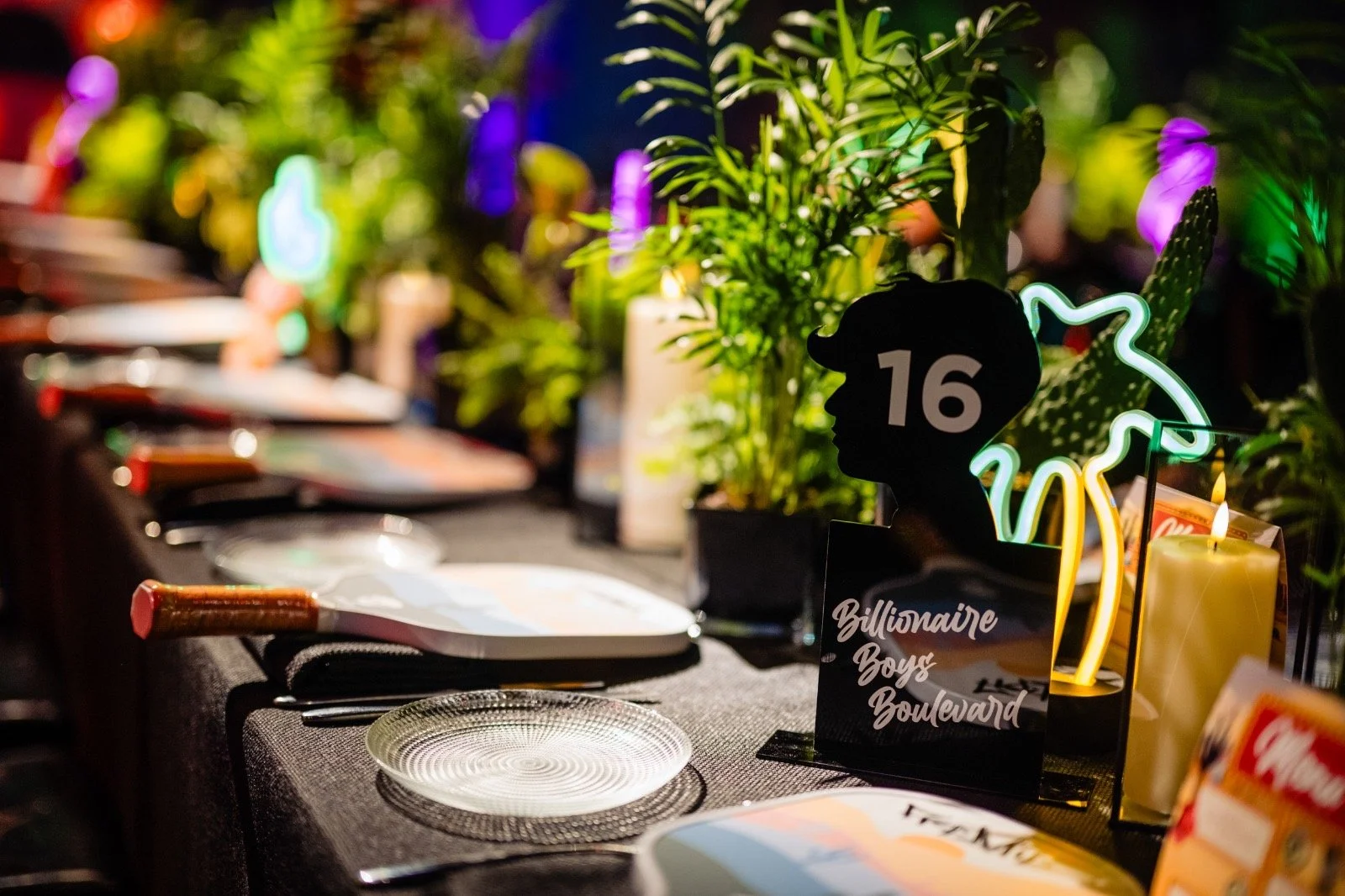 A table set for dining with plates and cutlery in front of a colorful, illuminated decorative sign showing a silhouette of a person's head with the number 16 inside, surrounded by green plants and candles, at a stylish event or celebration.