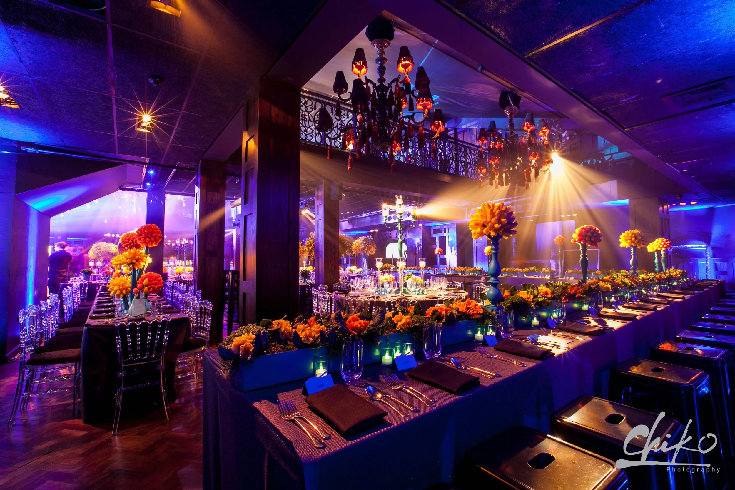 Elegant event hall with long banquet tables decorated with yellow and orange flower centerpieces, set with silverware, glasses, and black napkins. The room is illuminated with colorful blue and purple lighting, featuring a grand chandelier hanging fr