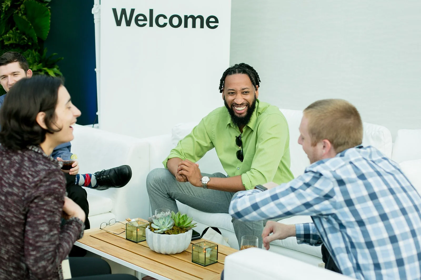 Group of five people sitting on white sofas, engaging in conversation, in a bright room with a 'Welcome' sign on the wall, a wooden coffee table with plant decorations and candles.