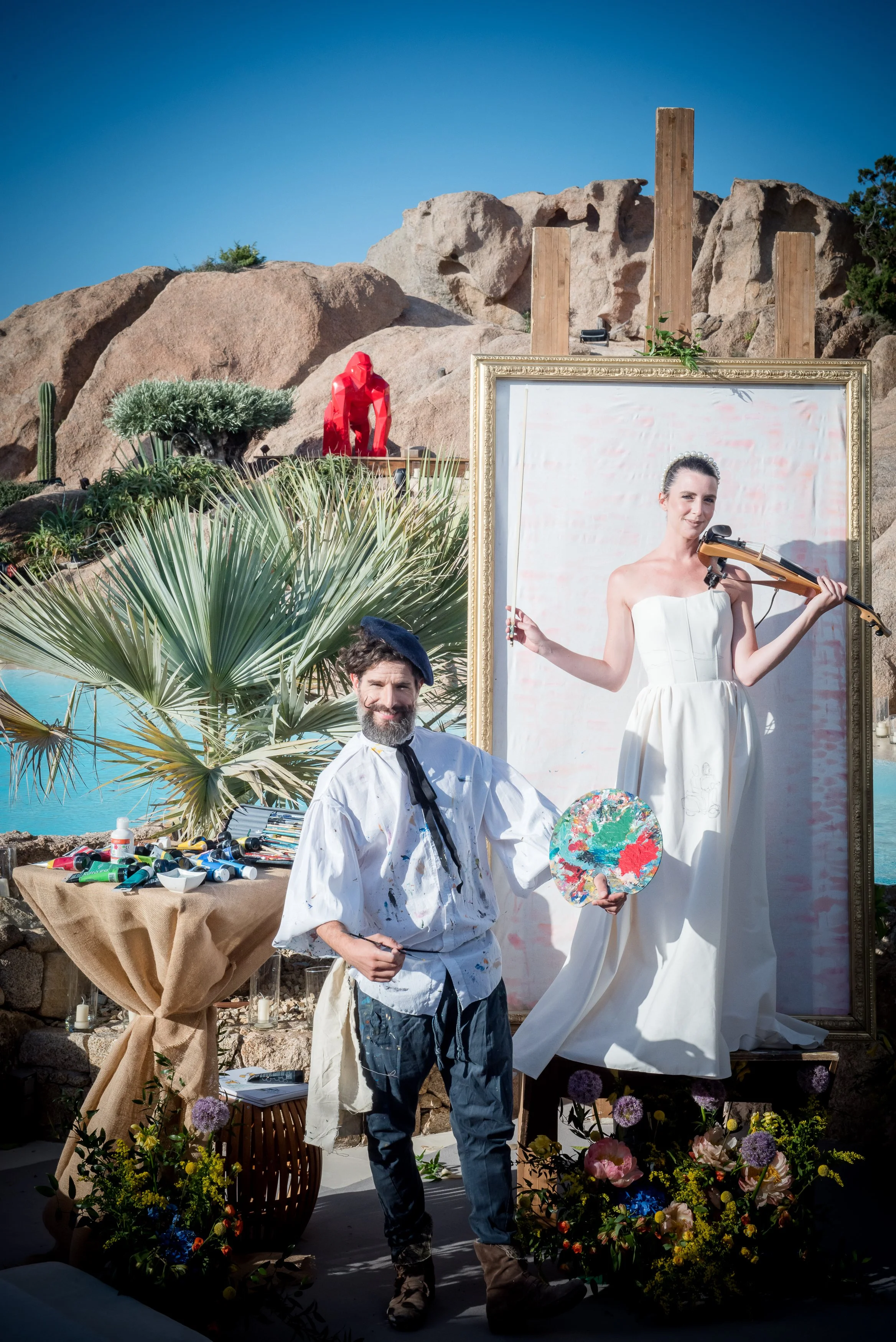 Two artists in an outdoor setting surrounded by flowers, plants, and art supplies. One is holding a paint palette and wears a painter's smock; the other is playing a violin and dressed in a white wedding gown, standing in front of a large framed canv