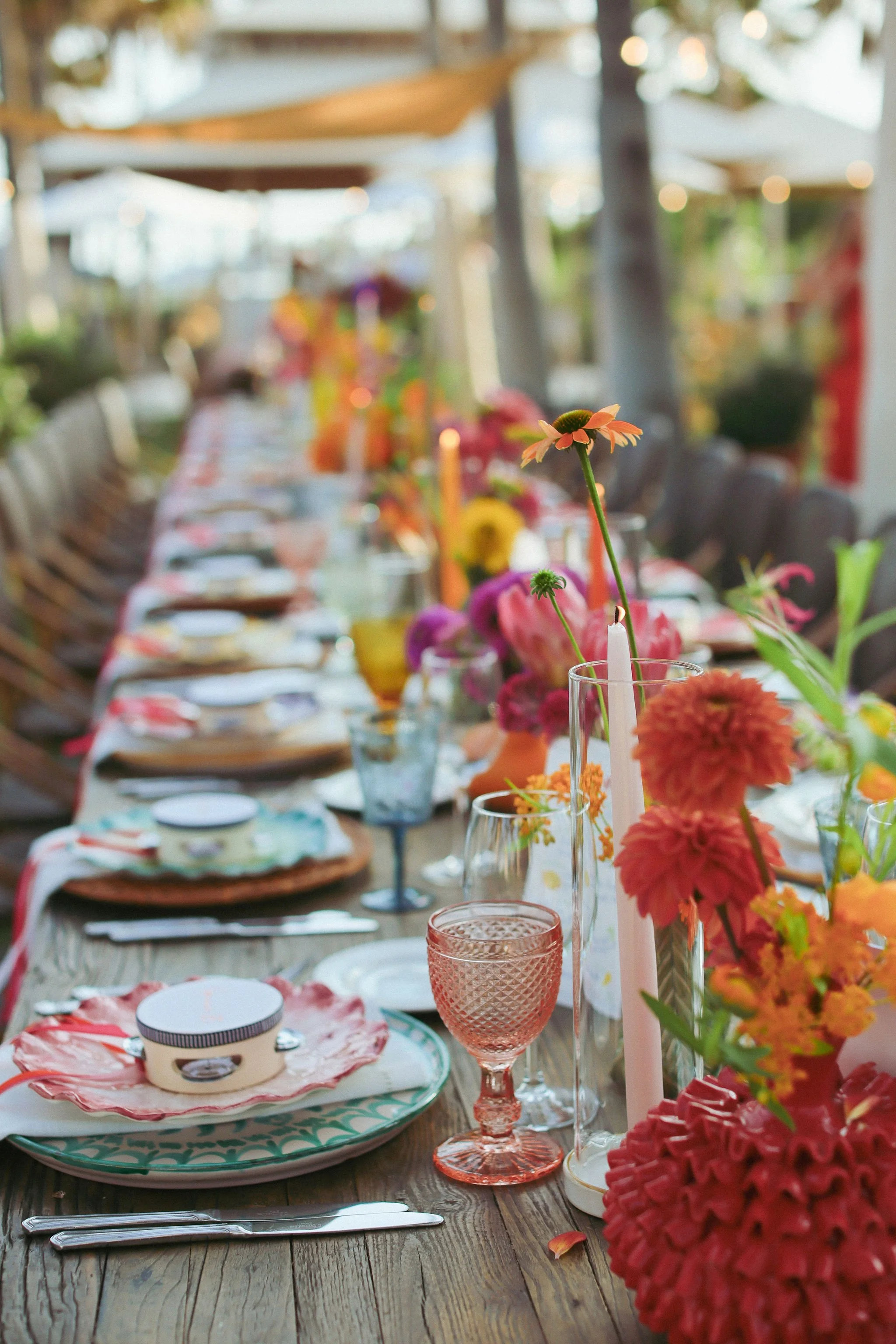 A table set for a celebration with colorful flowers in vases, pink taper candles, glassware, and plates with patterned napkins, on a wooden surface in an outdoor setting with blurred background of trees and umbrellas at. Anantara Villa Padierna Resor