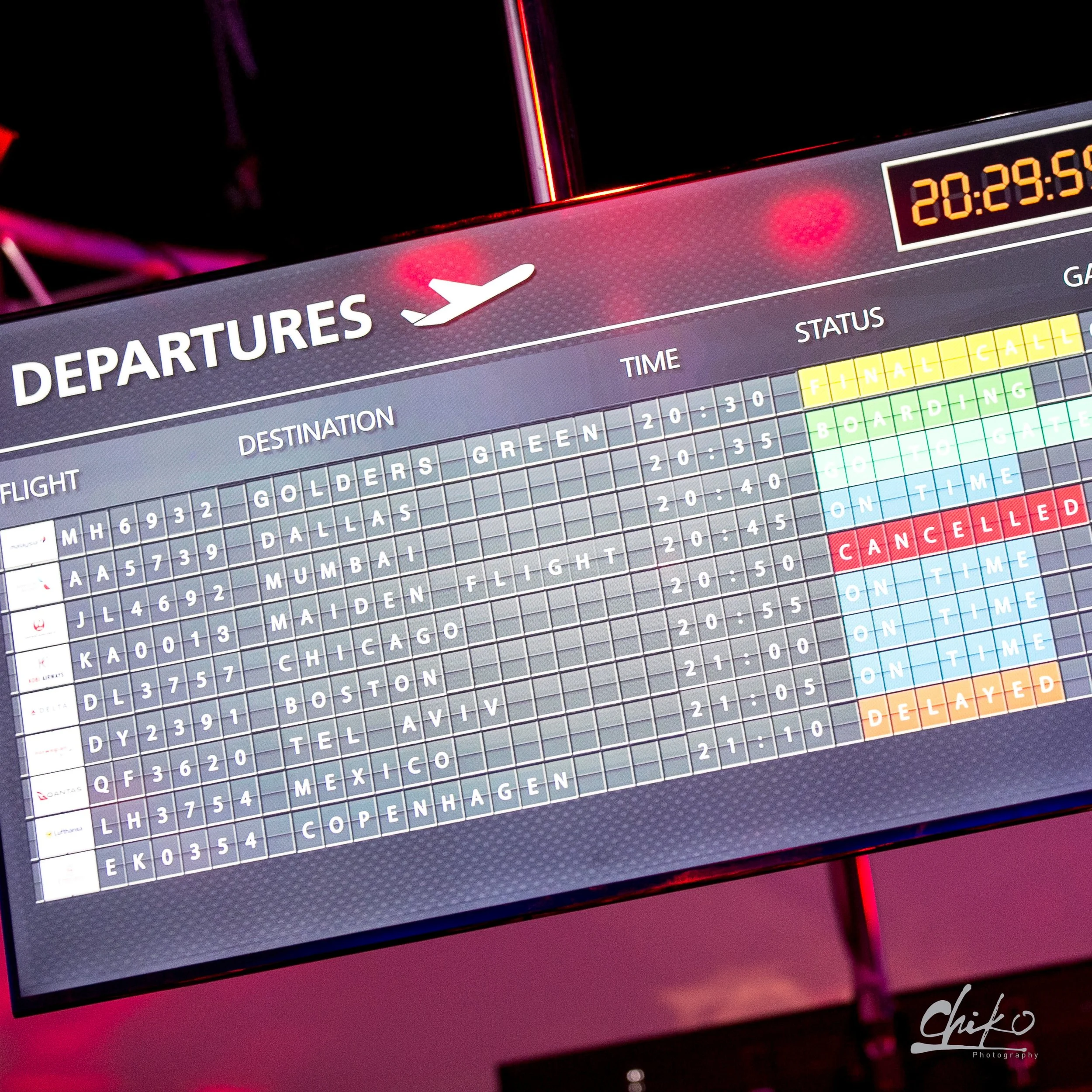 Photo of a digital departure board at an airport displaying flight information, with a background illuminated in pink and red lighting.