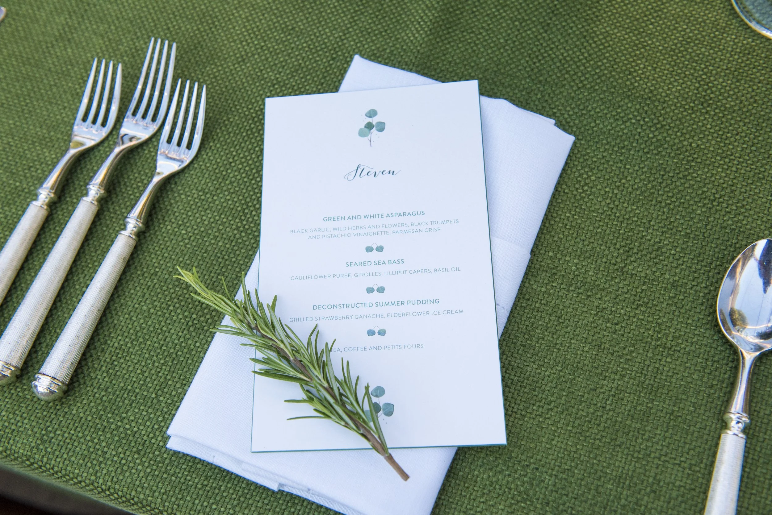 A wedding table setting with a green textured tablecloth, three forks on the left, a white napkin, a menu card with a green and white theme and a sprig of rosemary, and a silver spoon on the right.