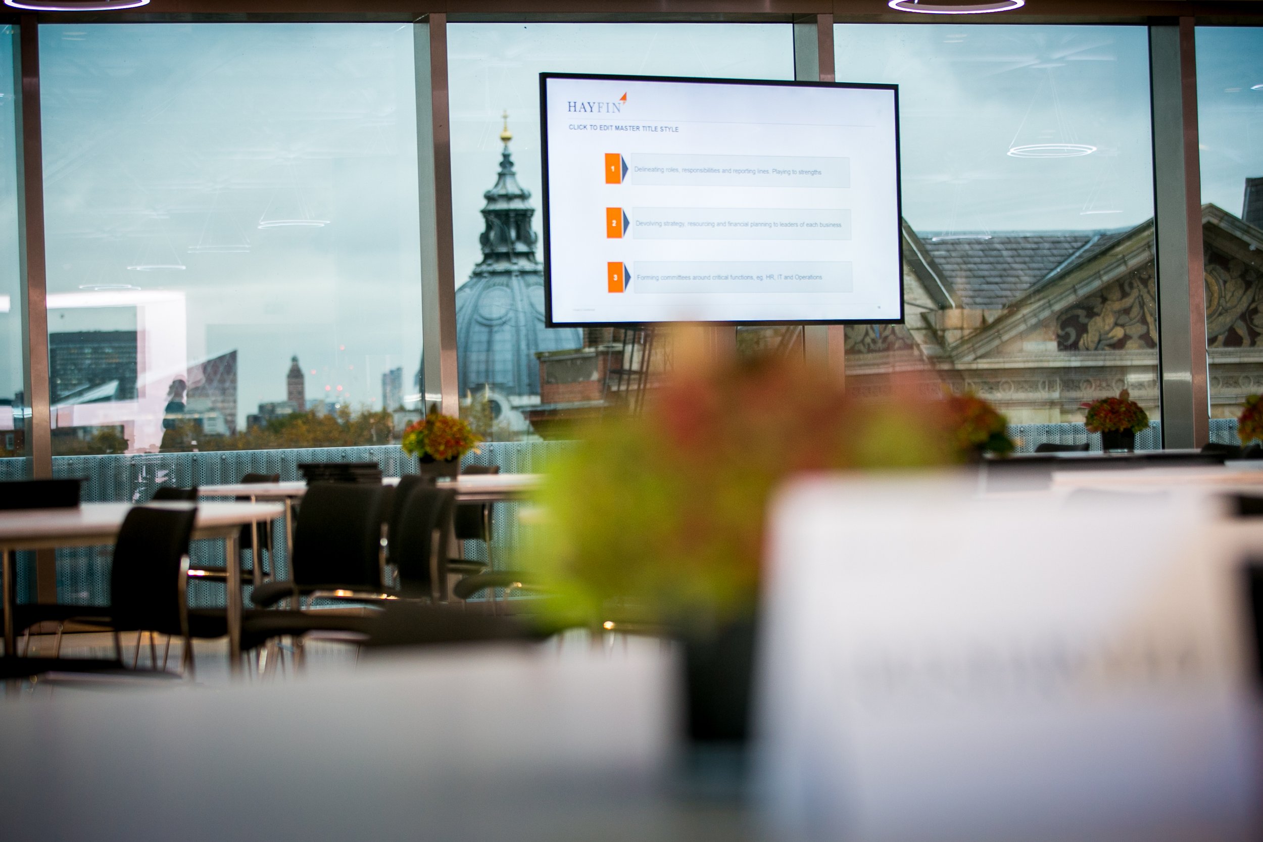 View of a modern conference room with large windows showing a cityscape and historic buildings. There are empty chairs and tables, and a large screen displaying a presentation titled 'Hayfin' with bullet points.