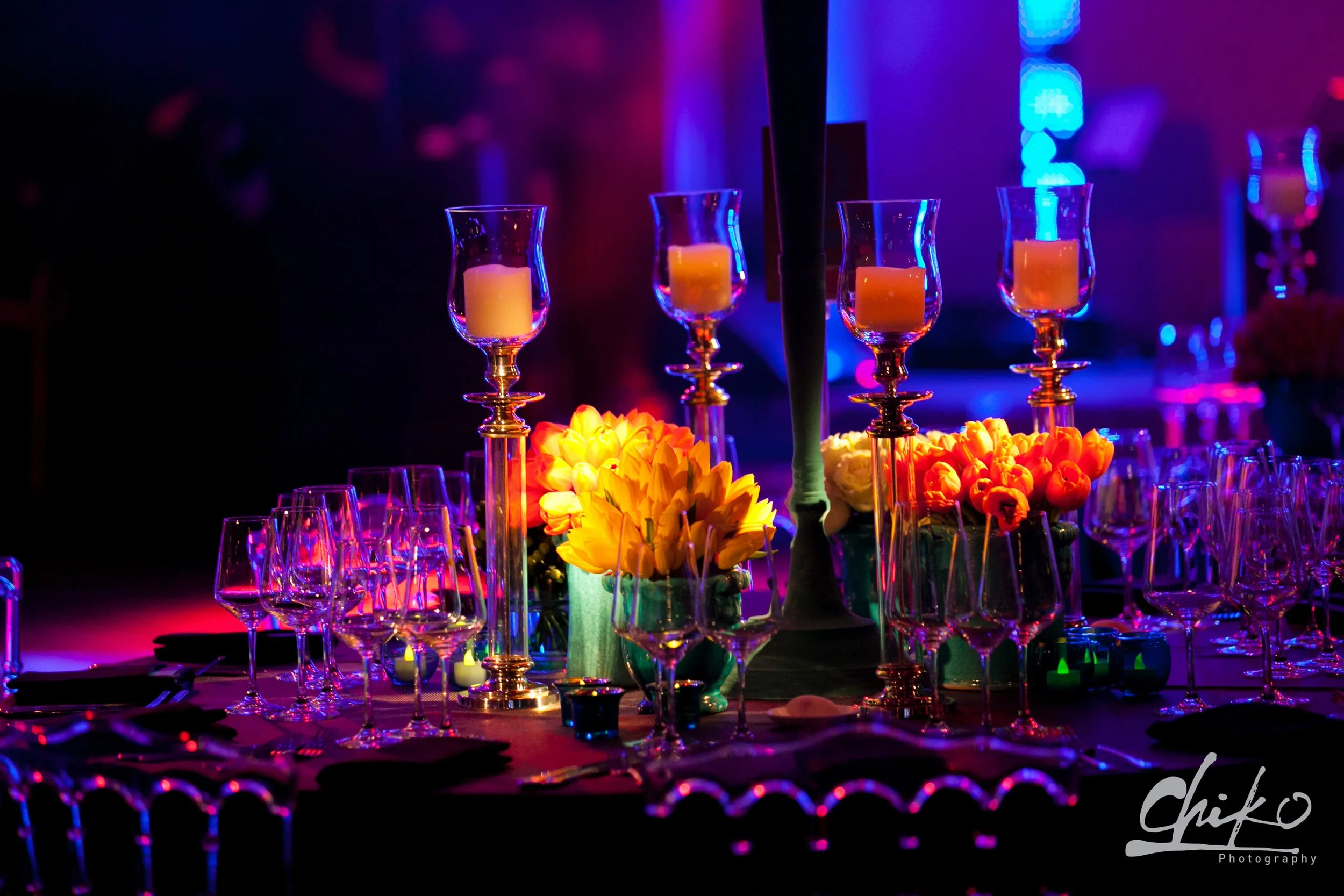 A table decorated with flower arrangements, candles in glass holders, and wine glasses under colorful ambient lighting at an event or celebration.