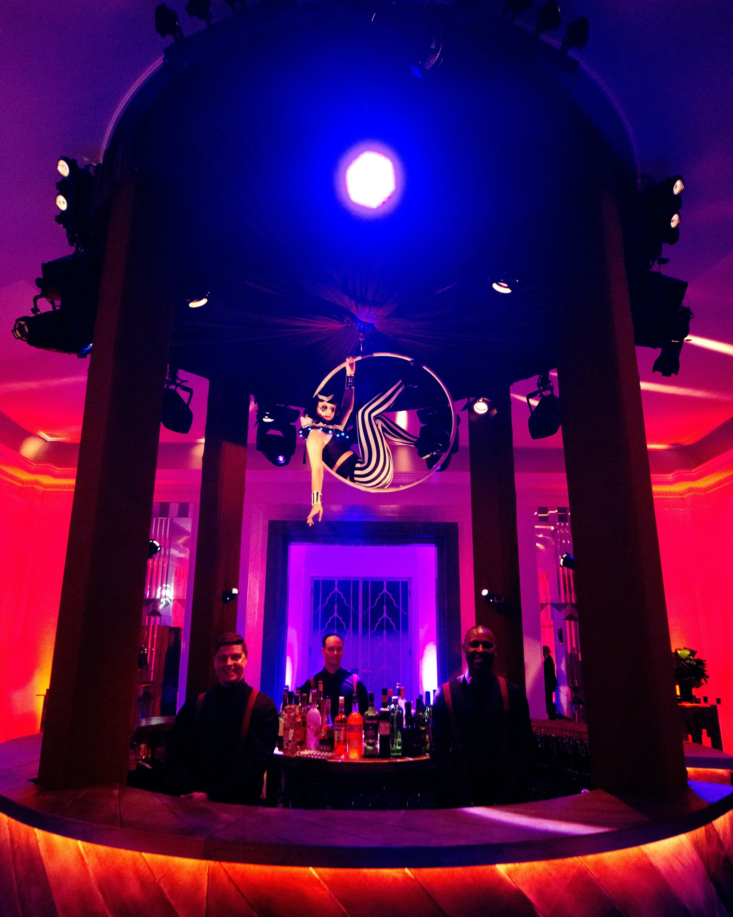 A bar with a colorful illuminated backdrop, a bartender behind the counter, and three people standing in front of the bar, in a dimly lit space with purple and red lighting.