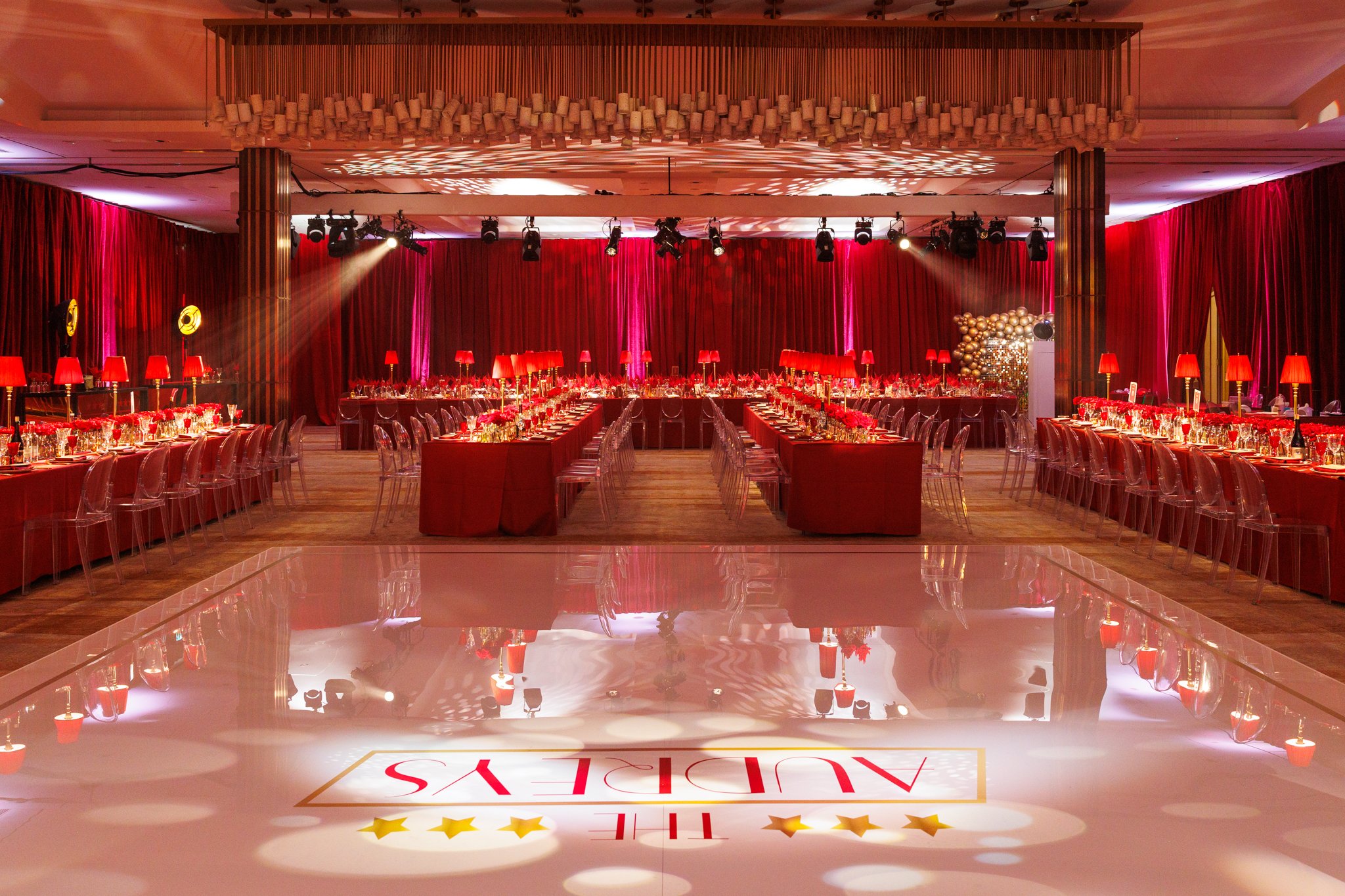 An event hall decorated with red curtains, tables with red tablecloths, red lamps, and floral centerpieces. Stage lighting and a balloon decoration are visible, with a dance floor in the foreground showing part of a logo or sign at The Grove 