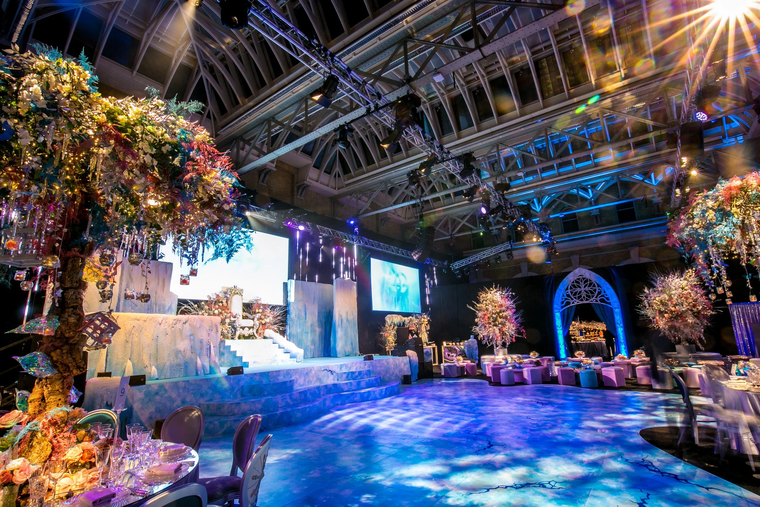 Elegant event venue with floral decorations, a decorated stage, and colorful lighting ICE PALACE BAT MITZVAH at Old Billingsgate 