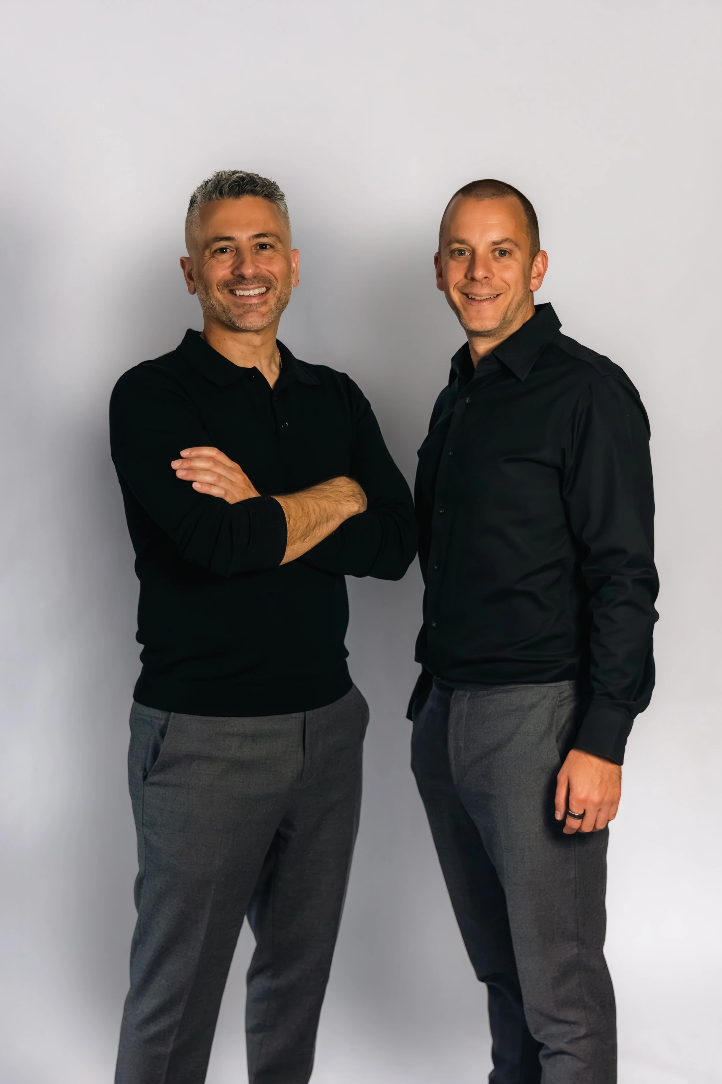 Laurie Weitzkorn and Stas Anastasiou standing side by side against a plain background, smiling at the camera, dressed in dark shirts and gray pants.