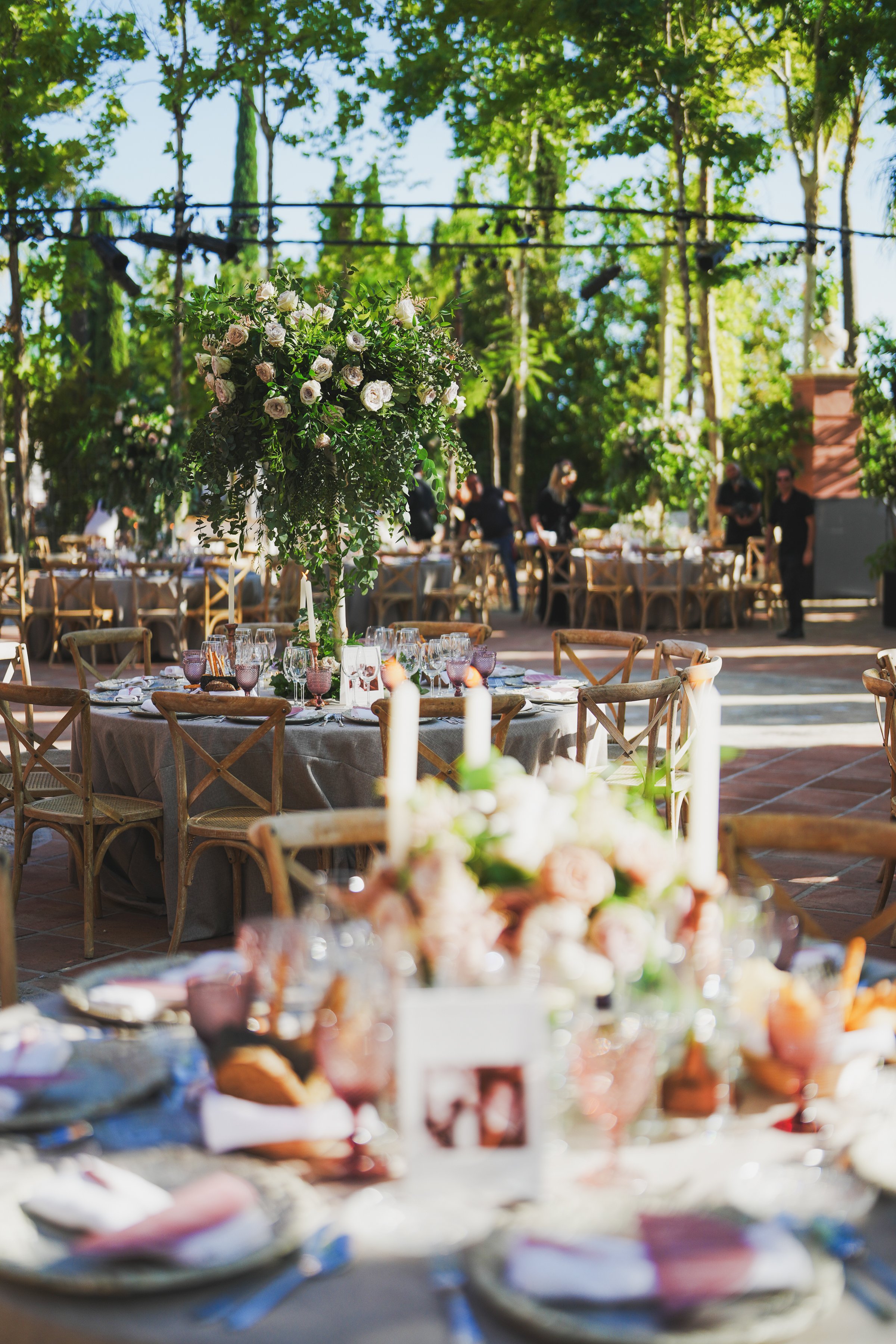 Outdoor wedding recpetion with decorated tables, floral centerpiece, and people in the background, surrounded by lush green trees.