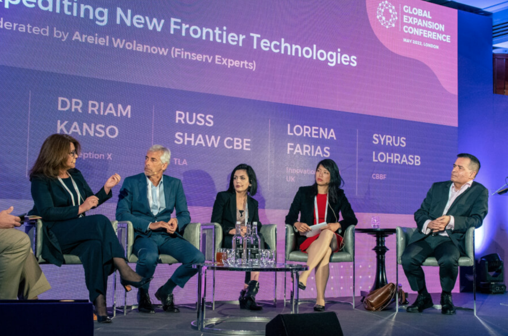 A panel discussion at the Global Expansion Conference in London, May 2022. Five speakers sit on stage with a large purple screen behind them displaying their names and affiliations. Six glasses of water and a pitcher are on a glass table in front of them.