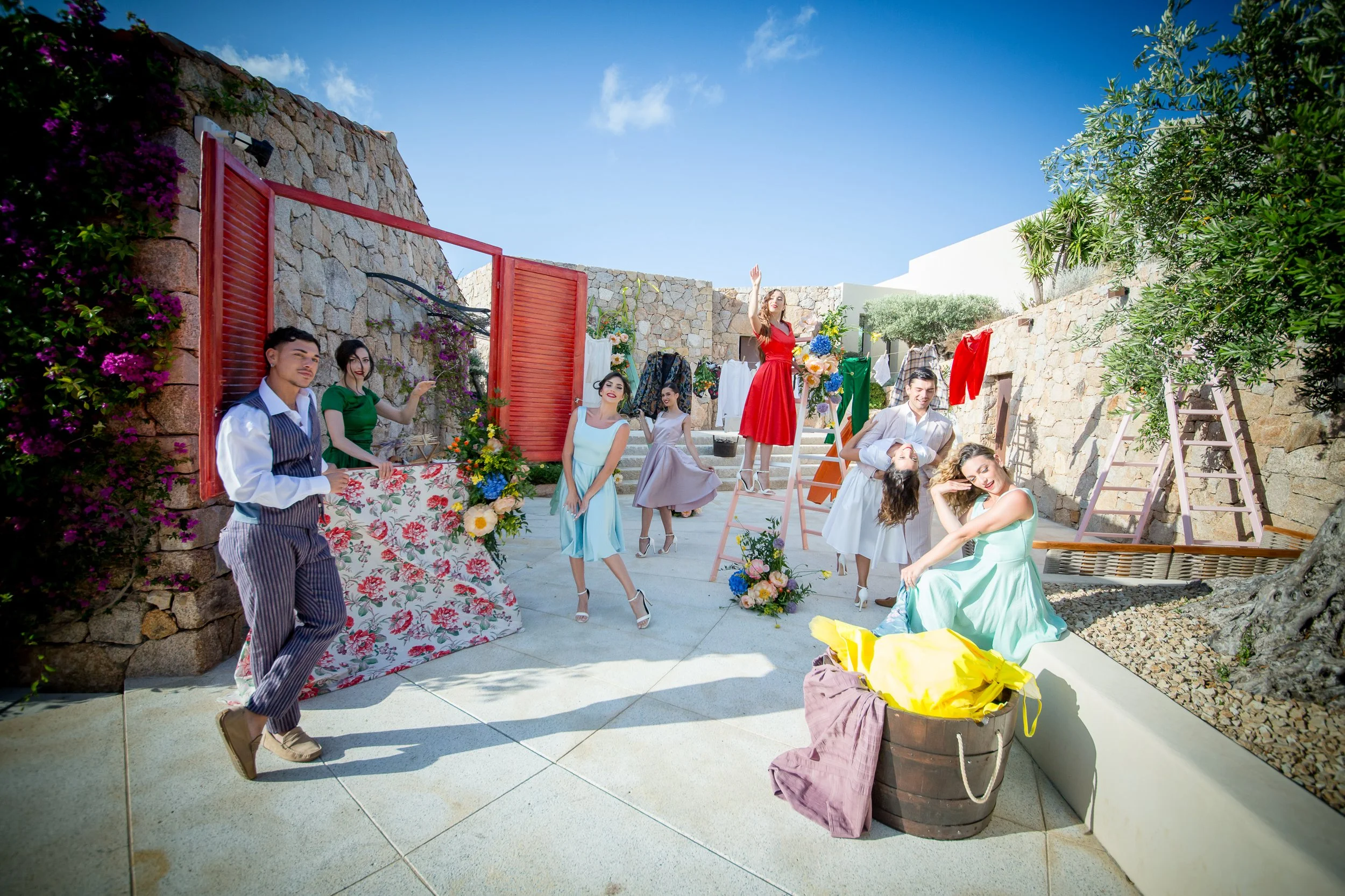 A group of people are gathered outdoors on a sunny day, with some posing and others talking or'standing'near a stone wall with flowers and laundry hanging. One woman in a red dress is standing on a ladder, waving, while a man in a white shirt is hold