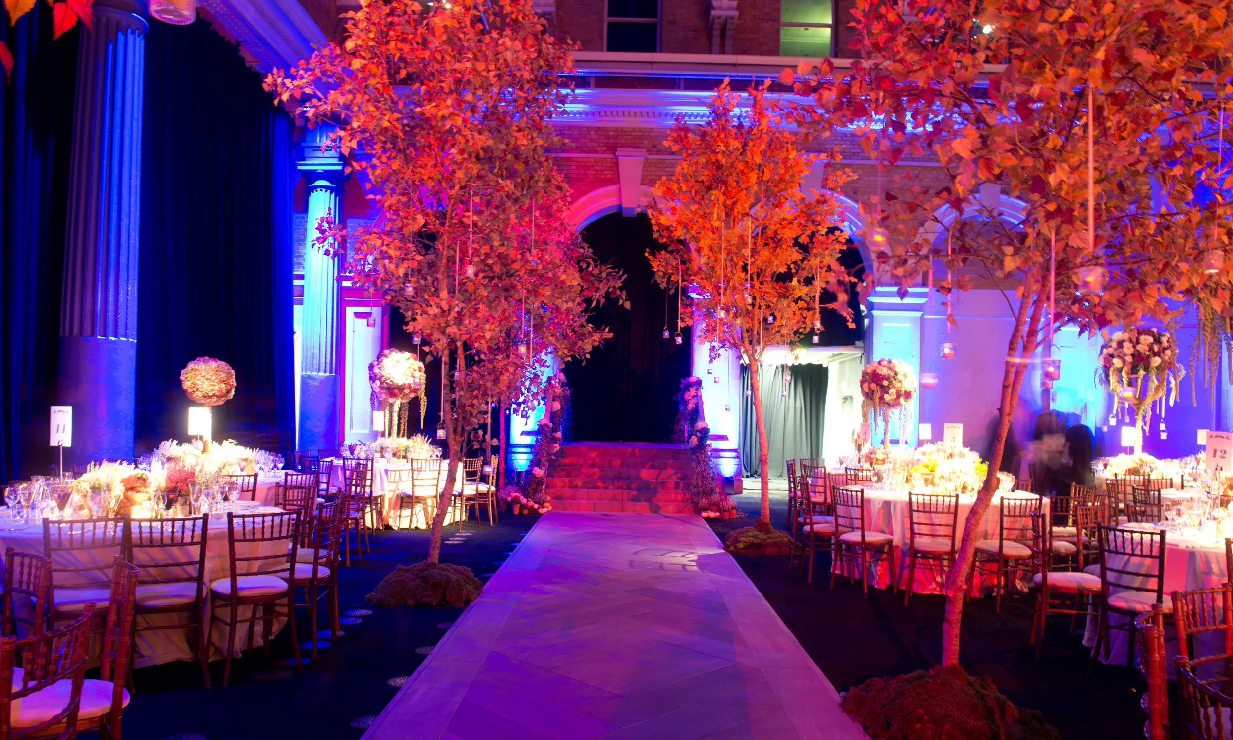 A decorated event space with a runway, round tables with chairs, and tall trees with red leaves illuminated by colorful lighting, set up for a celebration or party.