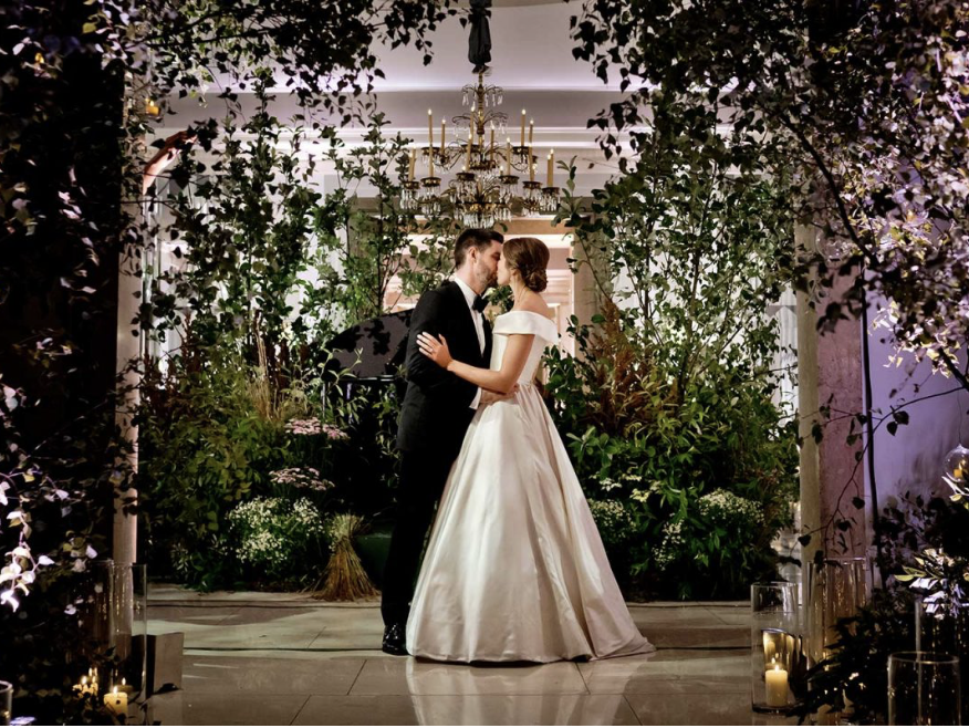 A bride and groom sharing a kiss in a decorated wedding setting with lush greenery and a chandelier overhead.