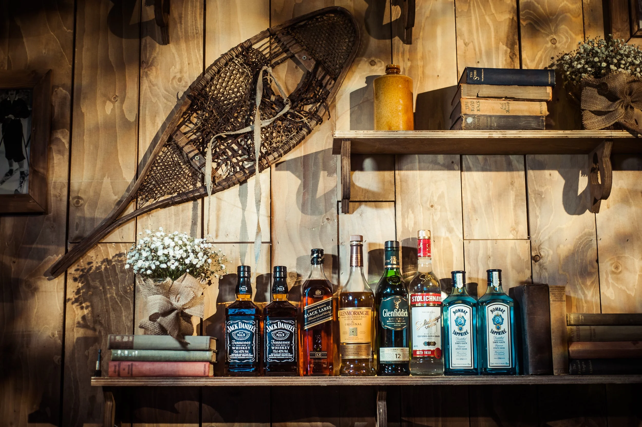 A wooden shelf wall with liquor bottles, a wicker sled, a yellow jar, books, a bouquet of white flowers, and a few small bottles.