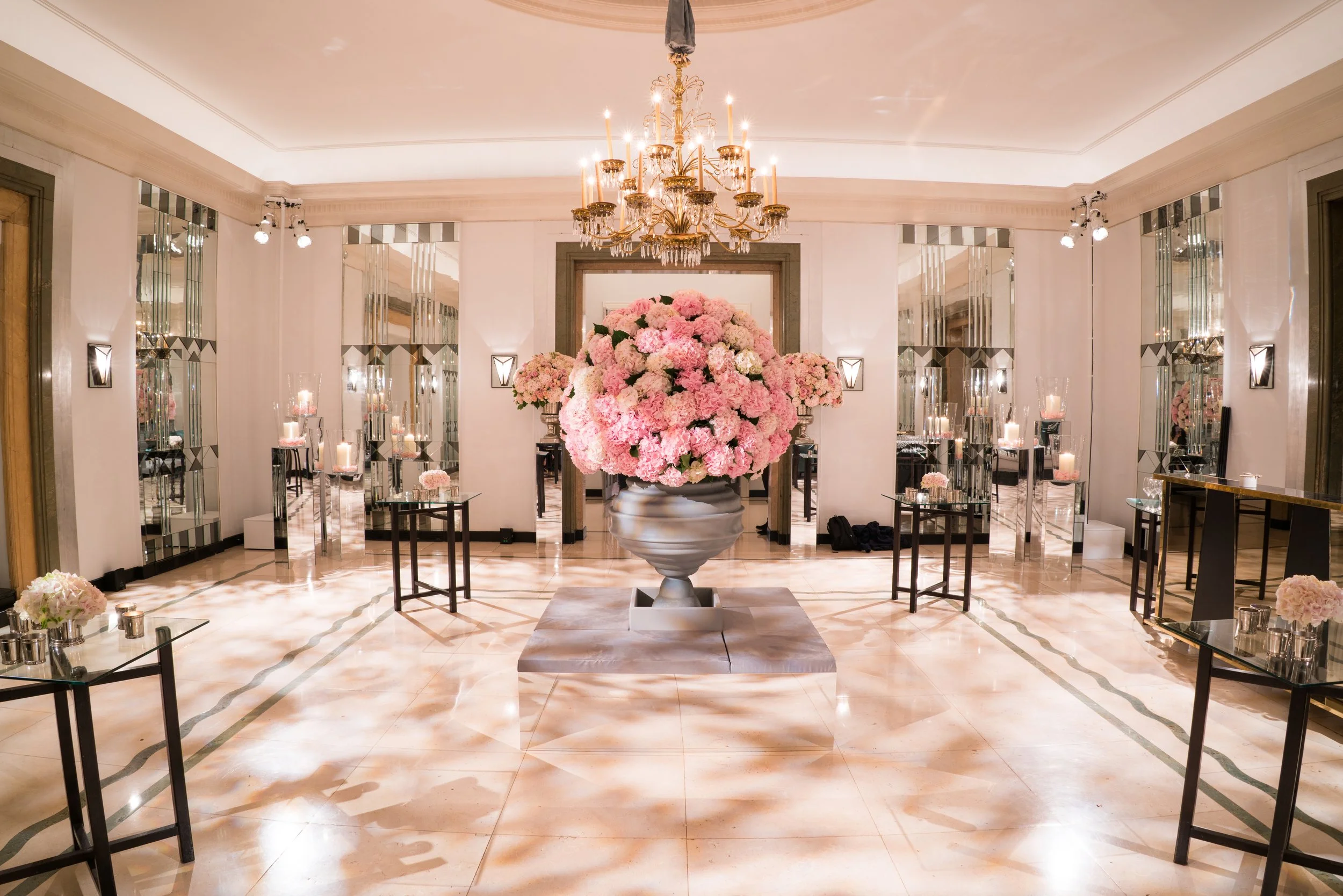 Elegant room decorated with a large pink hydrangea flower arrangement on a pedestal, a gold chandelier with candles, and mirrored wall panels reflecting candle displays, creating a luxurious ambiance.