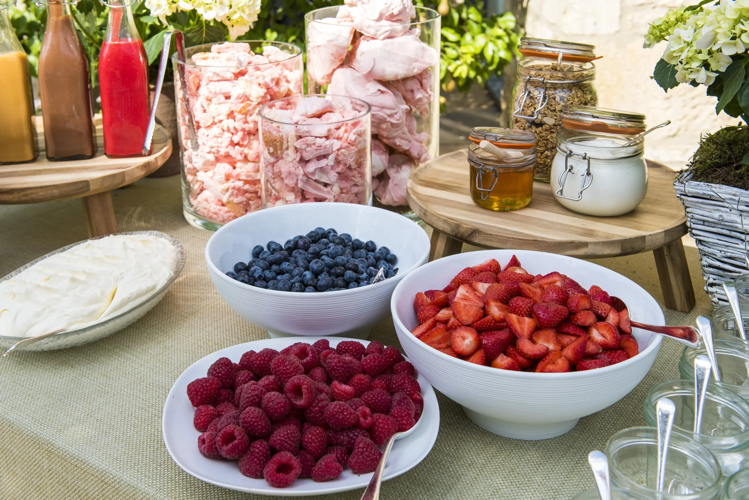 A variety of fresh berries including raspberries, strawberries, and blueberries in white bowls, along with containers of yogurt, honey, granola, and pink meringues on a table set outdoors.