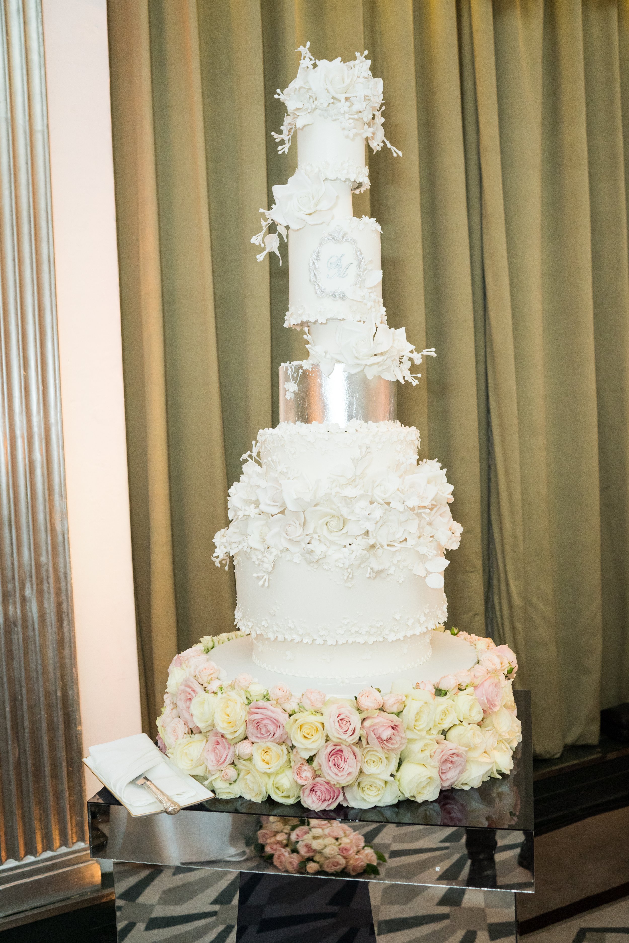 A tall wedding cake decorated with white and pink roses at the base, white flowers, and embossed designs, topped with a small floral arrangement, is displayed on a reflective black table with wedding utensils nearby inside a decorated indoor venue wi