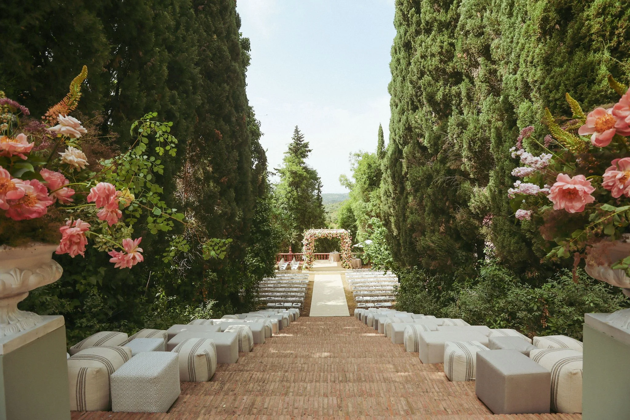 An outdoor wedding ceremony setup in a lush garden, with rows of white chairs on a brick pathway, flanked by tall, dense trees and decorated with flower arrangements, facing an archway covered in flowers and greenery at Anantara Villa Padierna Resort
