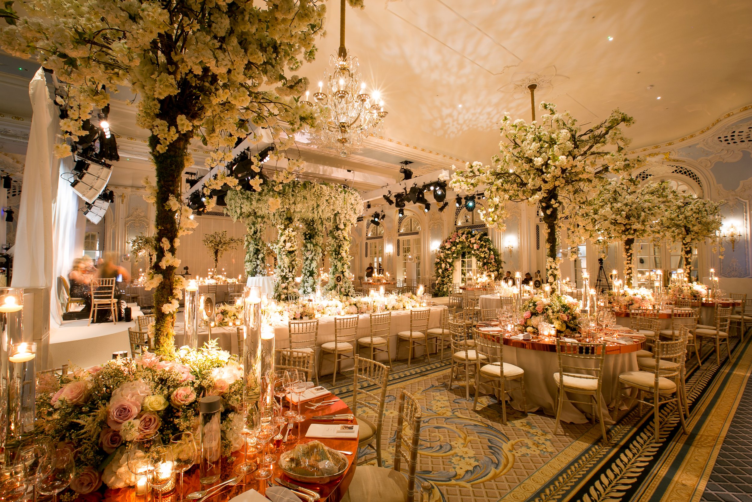 An elegant banquet hall decorated for a wedding with chandeliers, lush floral arrangements, and round tables set with candles, glasses, and tableware.