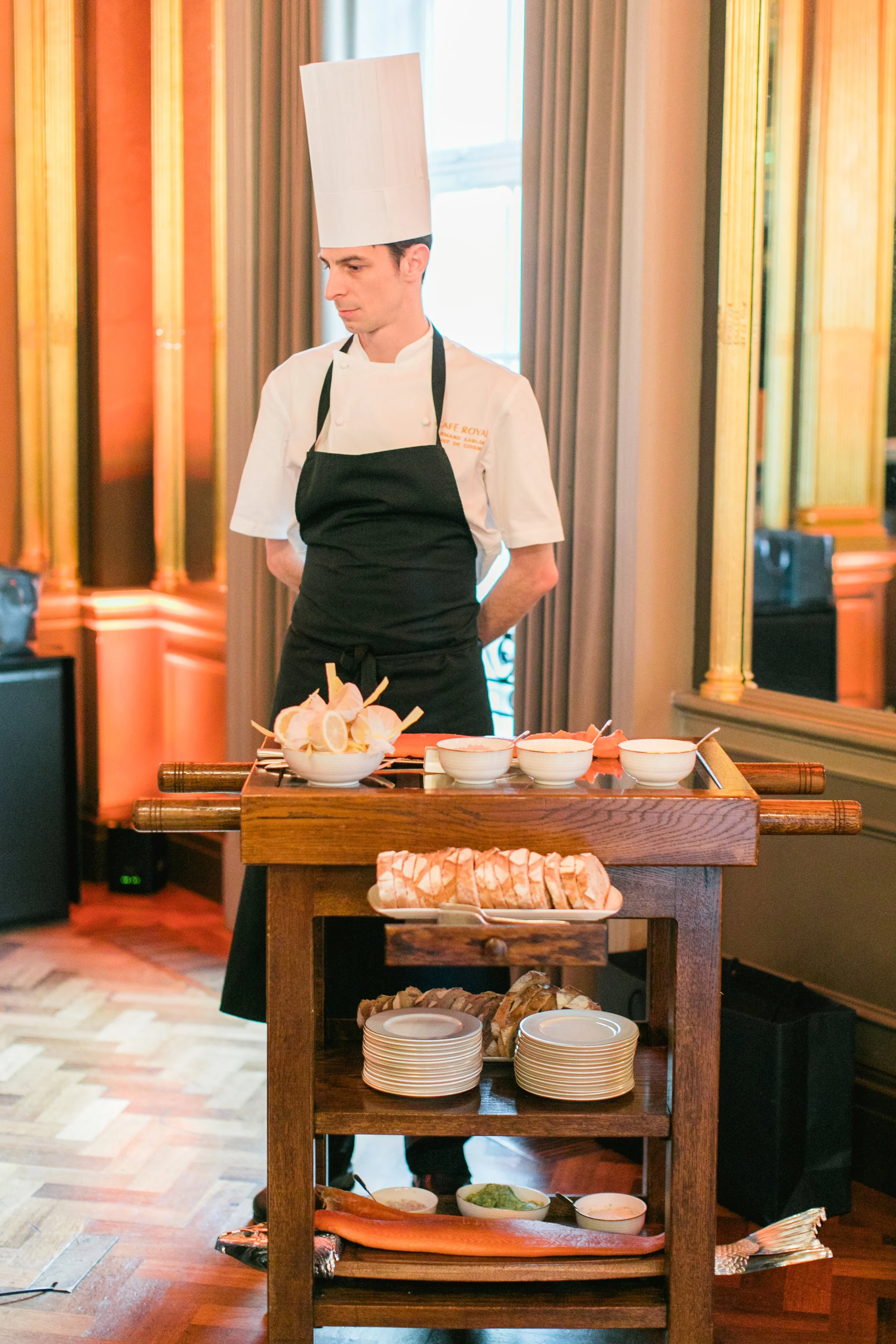 A chef wearing a tall white hat and white uniform with a black apron stands behind a wooden serving cart with various bowls of food and plates, in a room with wood flooring and curtains.
