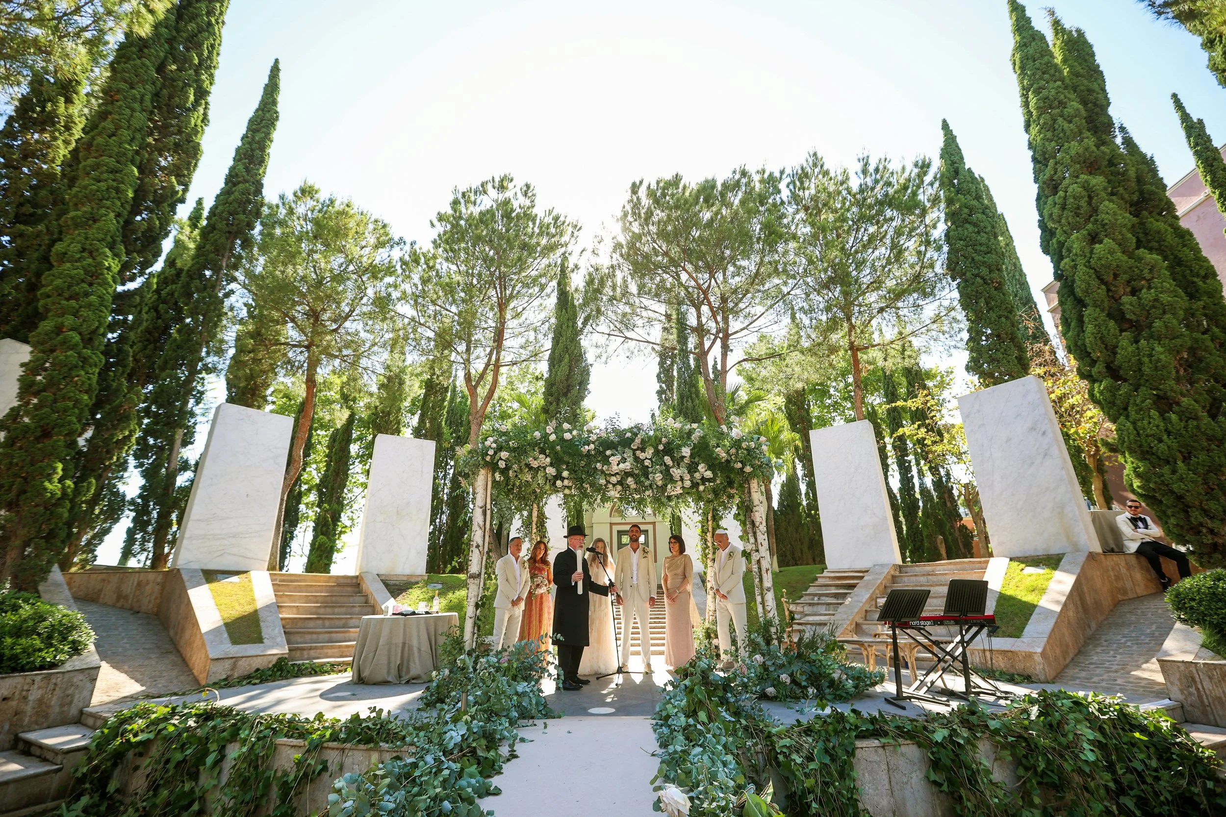 Wedding ceremony taking place outdoors surrounded by tall trees and lush greenery, with people dressed in formal attire under a floral arch.