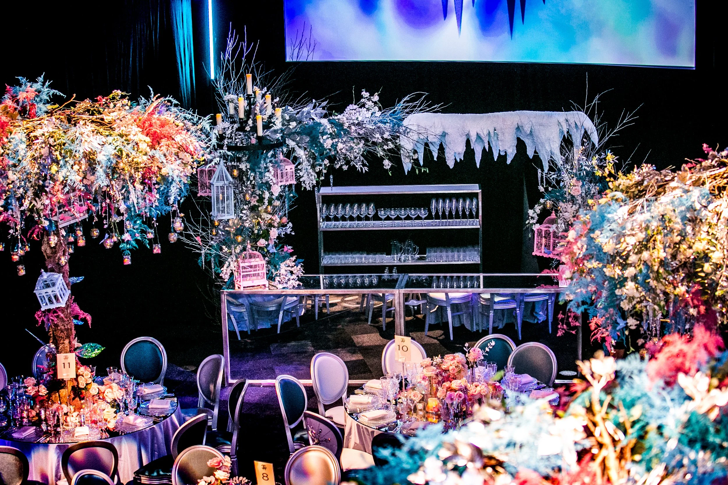 Interior of an event space decorated for a celebration with floral arrangements, fairy lights, and hanging lanterns on tables, with a stage and backdrop in the background. ICE PALACE BAT MITZVAH at Old Billingsgate 