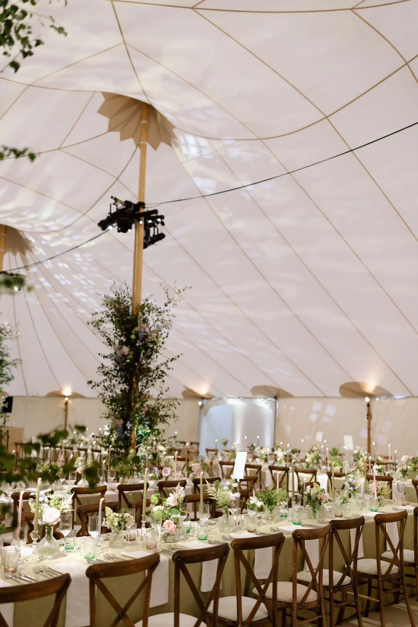 A decorated event space with long banquet tables arranged in a U-shape, adorned with flower arrangements, candles, and place settings. The ceiling is a large white fabric canopy with gold lines and a parasol hanging from the ceiling. There are string
