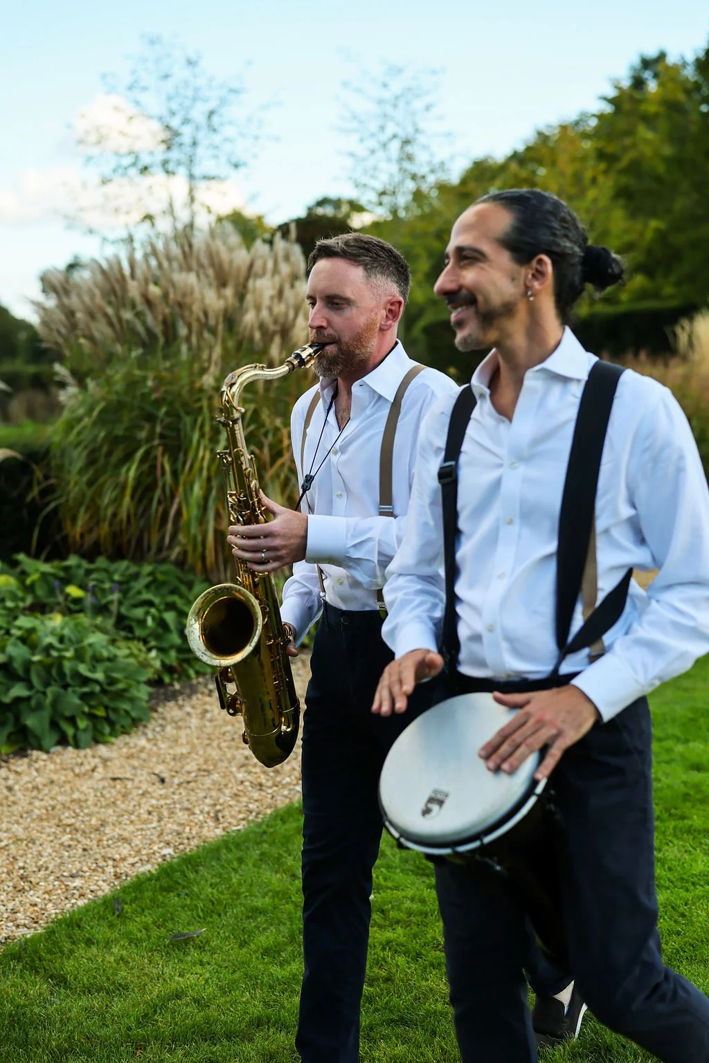Two men in white shirts and dark trousers walking outdoors; one playing a saxophone, the other holding a tambourine, with greenery and cloudy sky in the background.