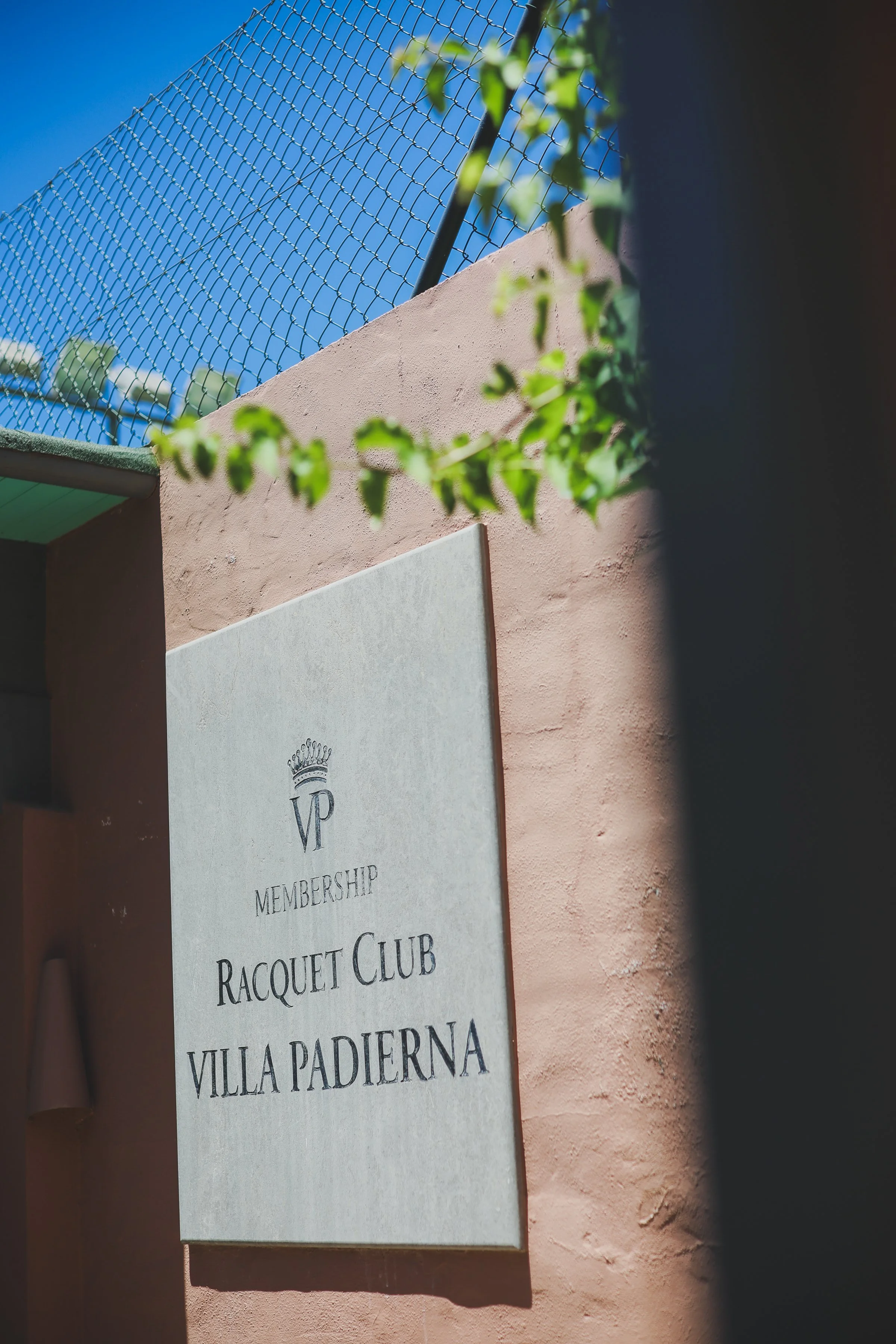 A white sign with black text and a crown logo, mounted on a pinkish wall, partially covered by green ivy, with a chain-link fence and a clear blue sky in the background.