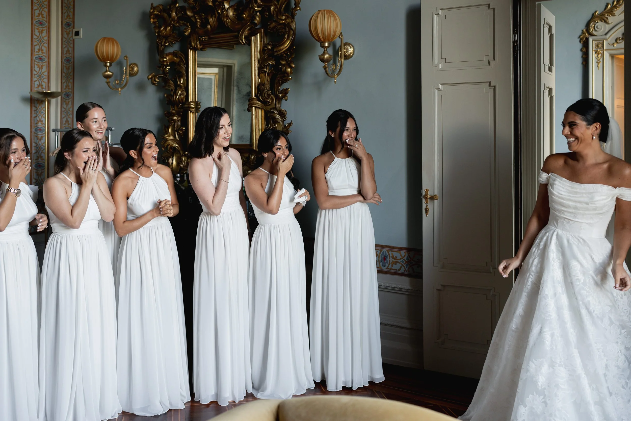Group of women in white dresses standing in a room, smiling and reacting to a woman in a wedding dress.