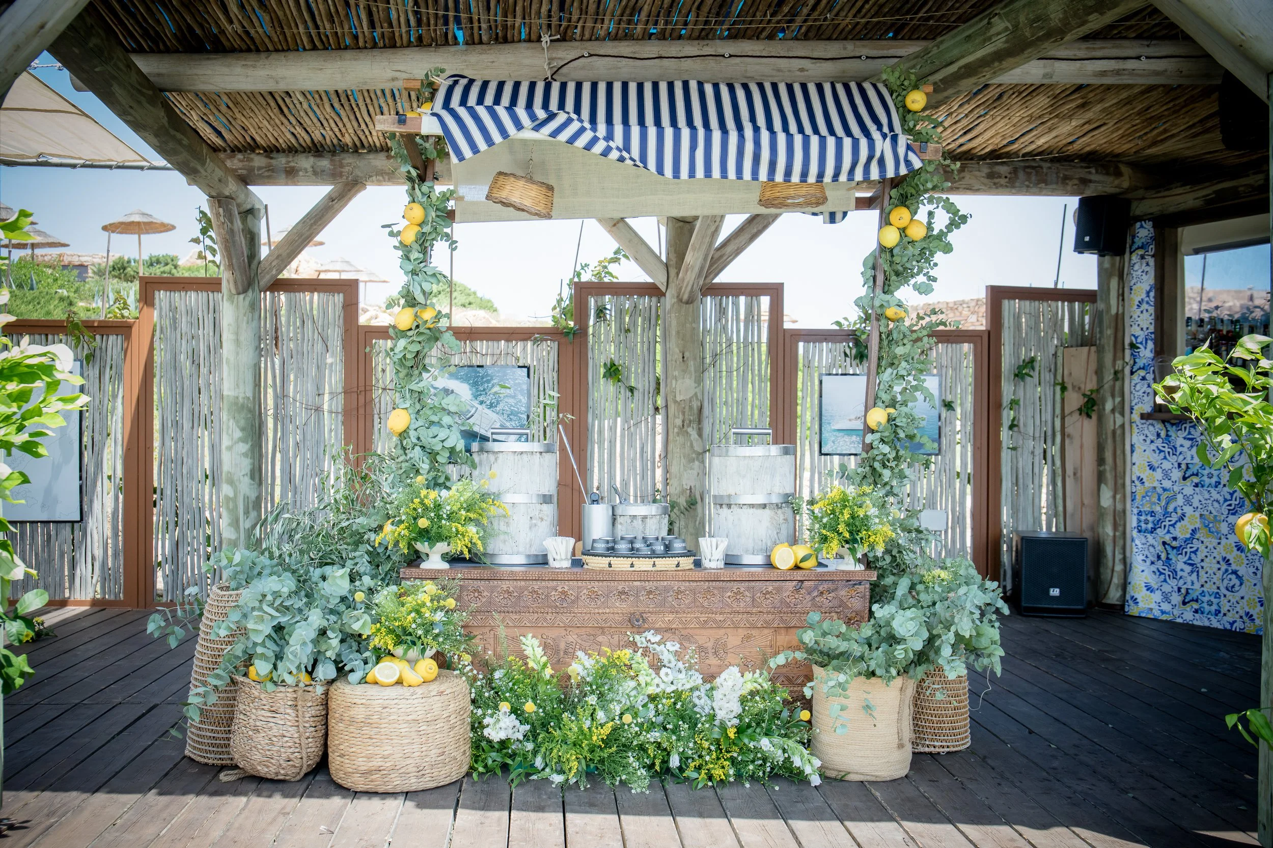A tropical outdoor bar decorated with lemon and green foliage, with a blue and white striped canopy, surrounded by wicker baskets, green plants, and yellow flowers, set on a wooden deck under a rustic roof.