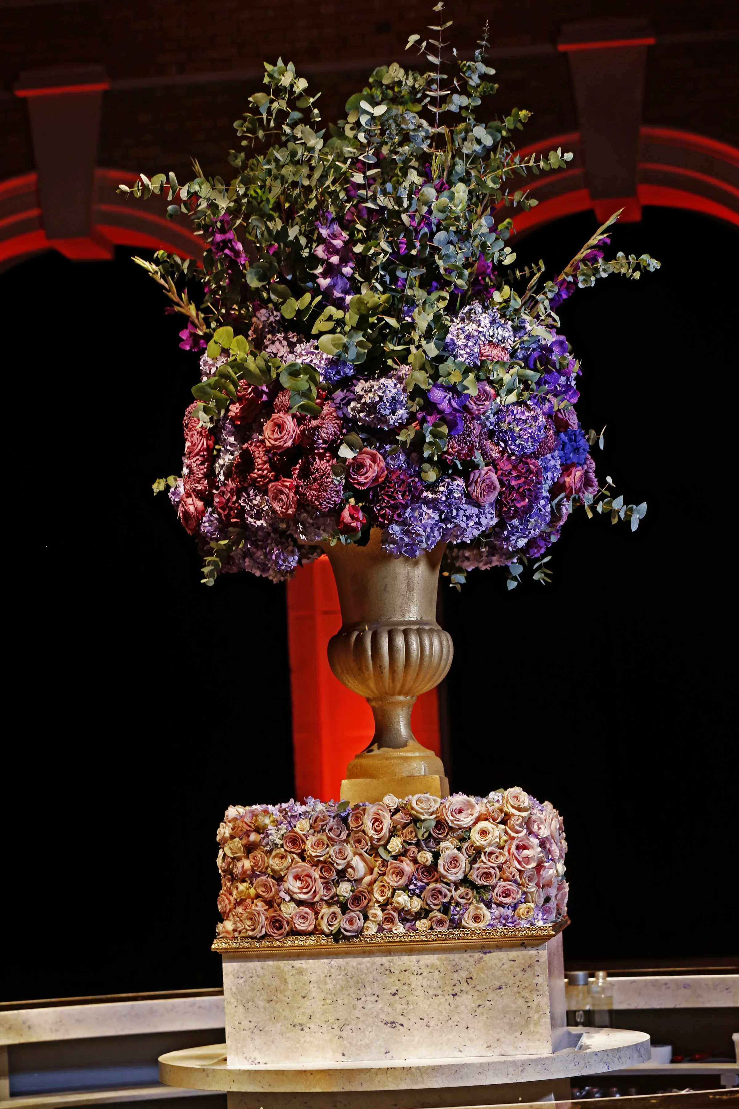 Two large multi-tiered flower arrangements with roses, hydrangeas, and greenery on marble bases, set against a dark background with red accents.